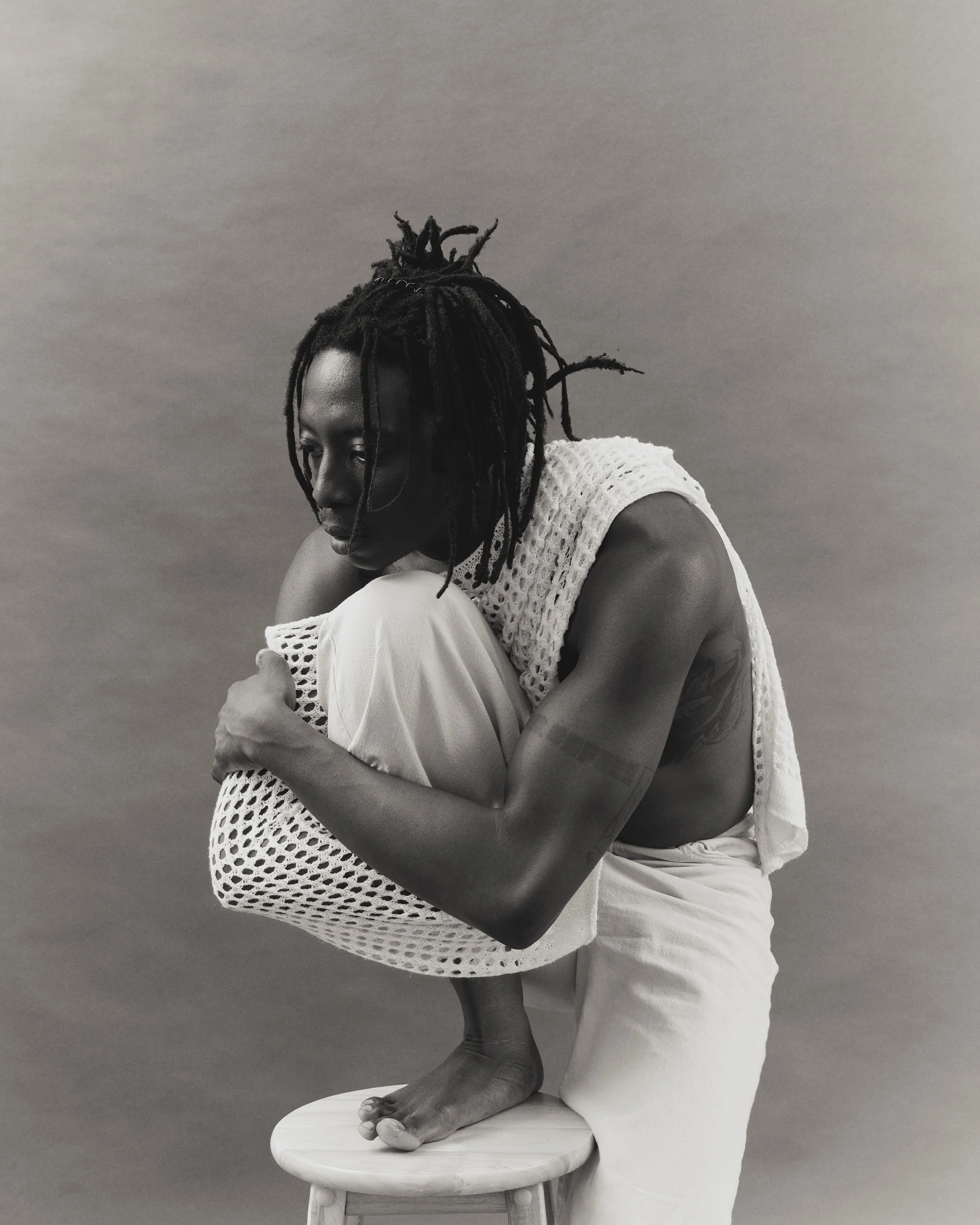 A person with dreadlocks crouches on a stool, hugging their knee, looking downward, dressed in light clothing, in a black and white portrait.