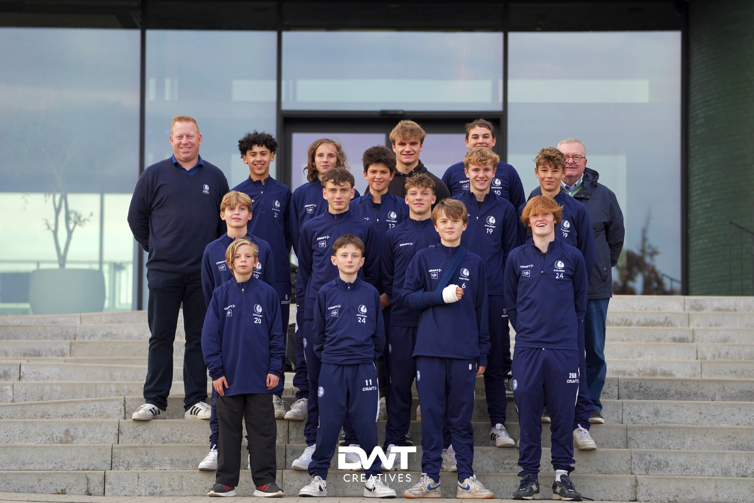 Groepsfoto van een sportteam met blauwe trainingspakken, poseren op een rij op een trap buiten bij een gebouw.