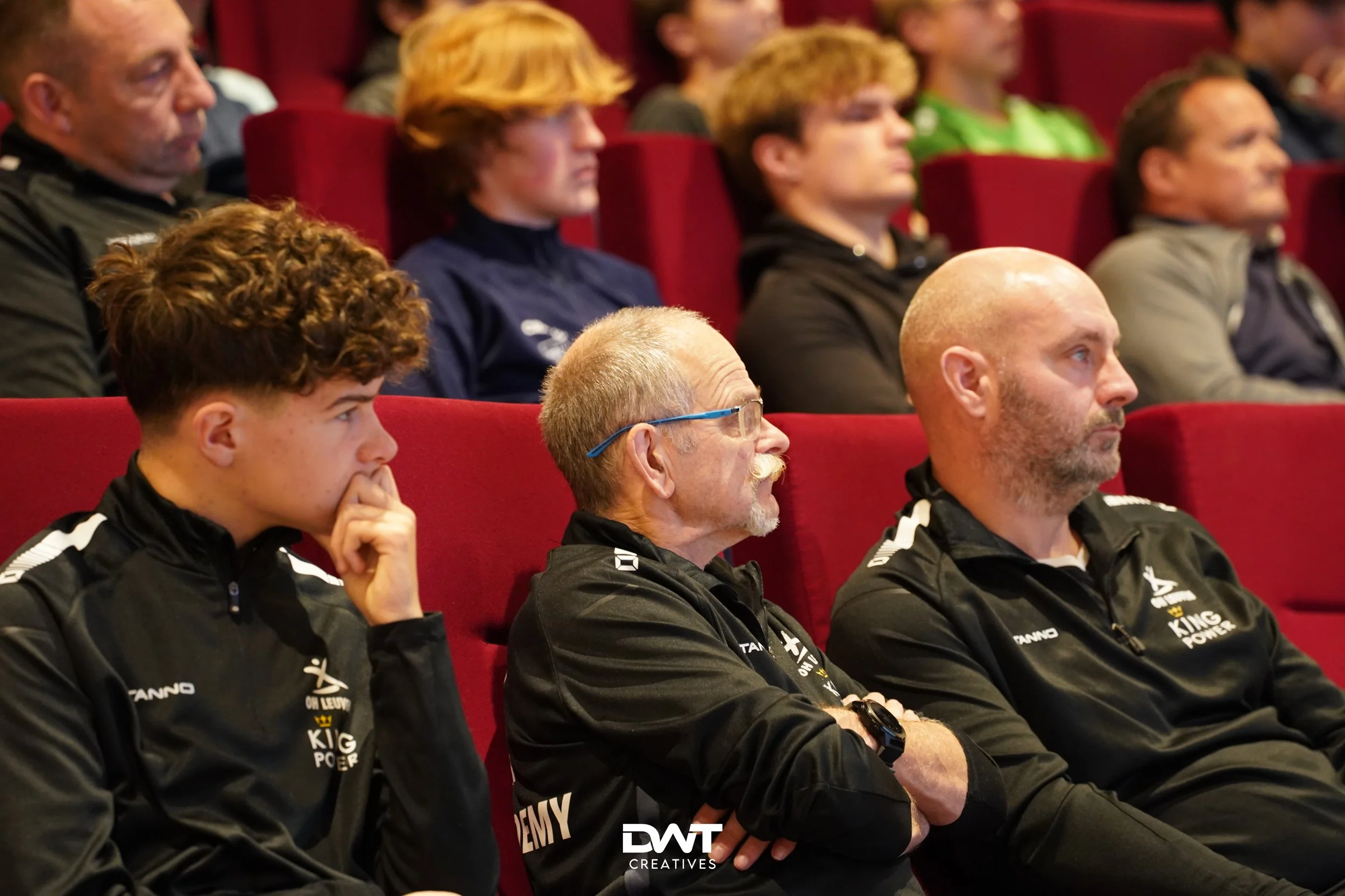 Mensen zitten in een zaal en luisteren aandachtig naar een presentatie. Ze dragen sportkleding met logo's van "Tanno" en "King Power". De stoelen zijn rood.