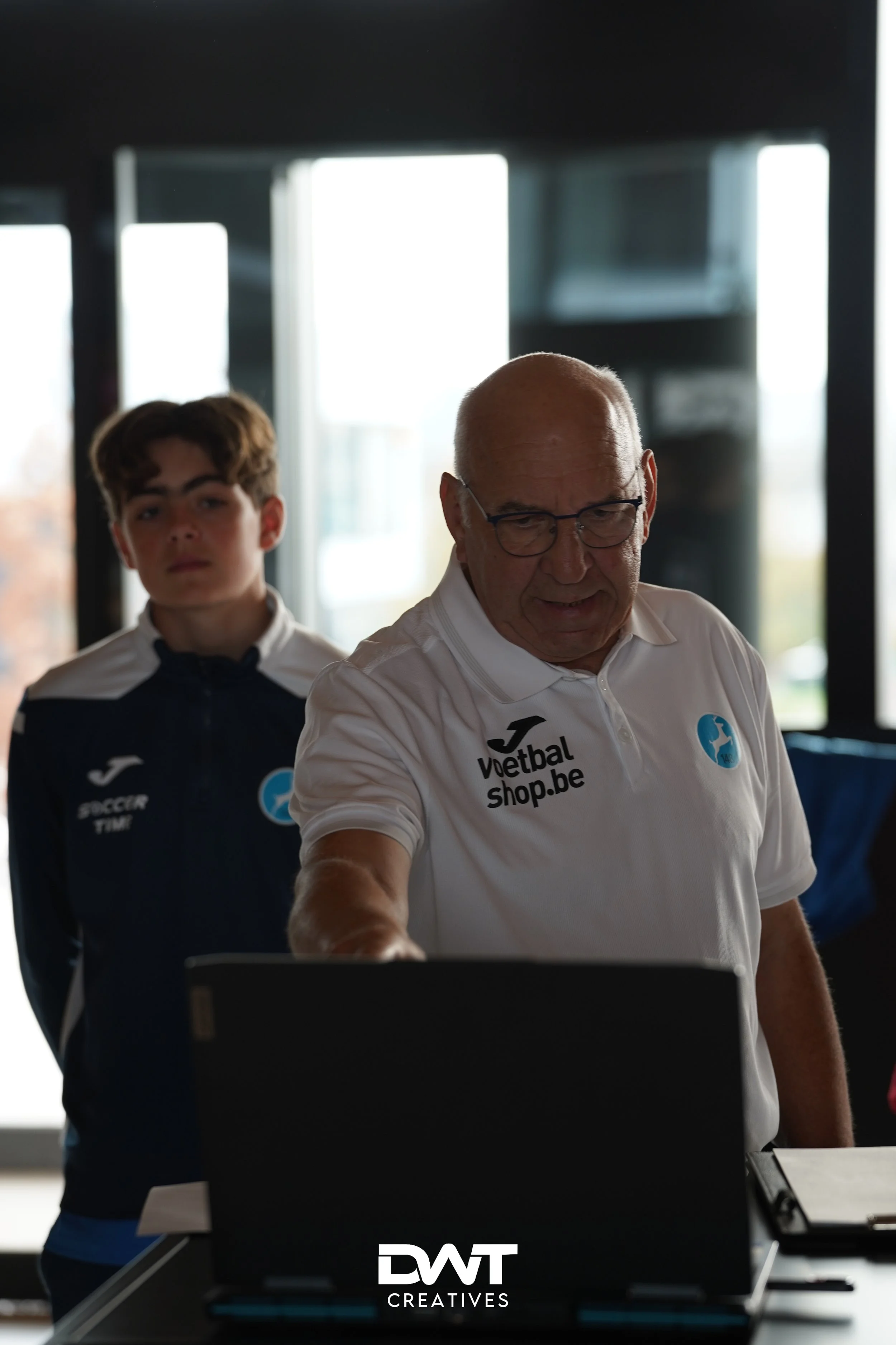 Twee mannen in sportkleding, een oudere die naar een computer wijst en een jongere op de achtergrond in een voetbalomgeving.