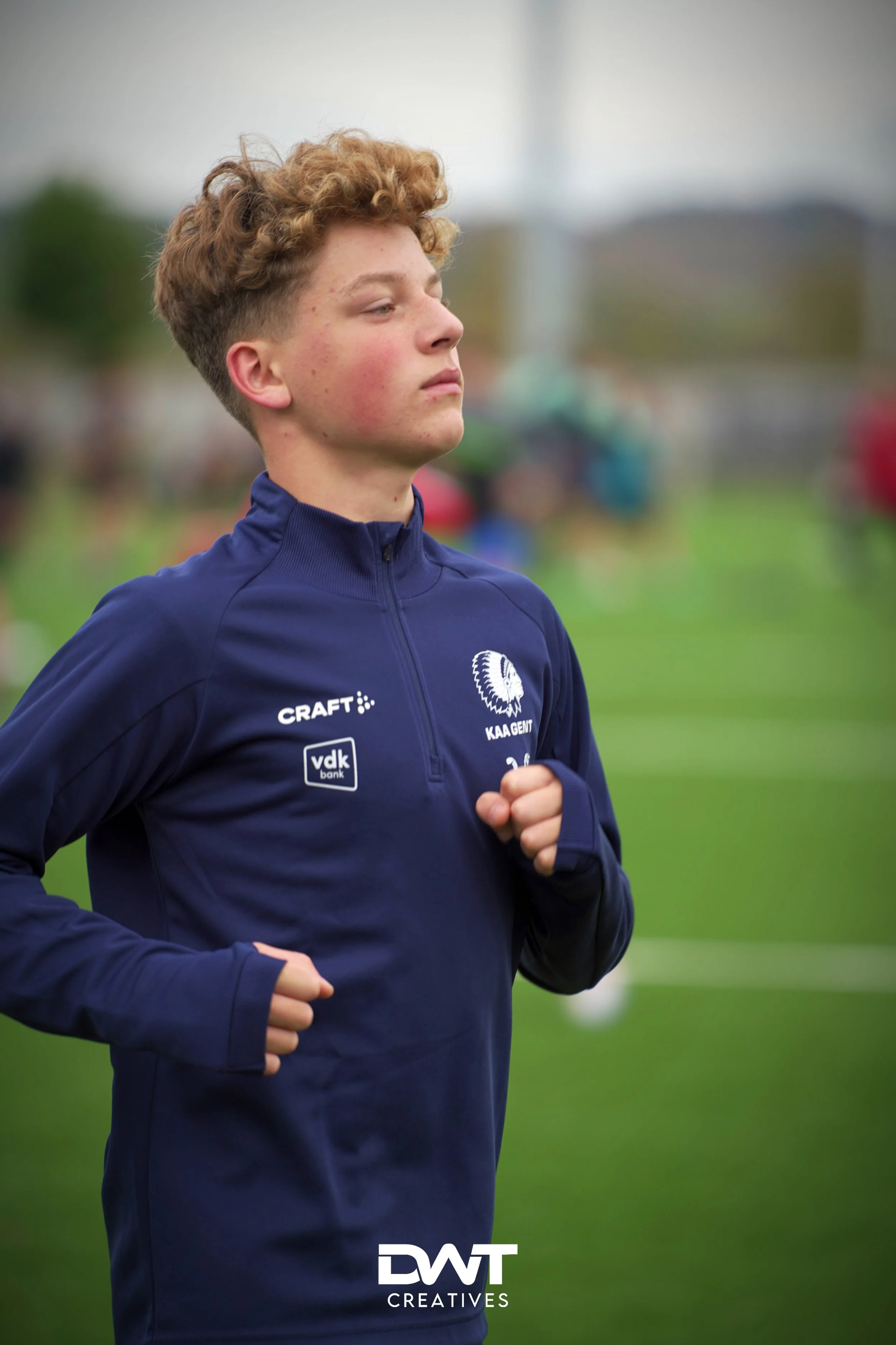 Jonge man in sportkleding van KAA Gent aan het joggen op een groene grasveld. Logo's van Craft en VDK Bank zijn zichtbaar op het shirt.