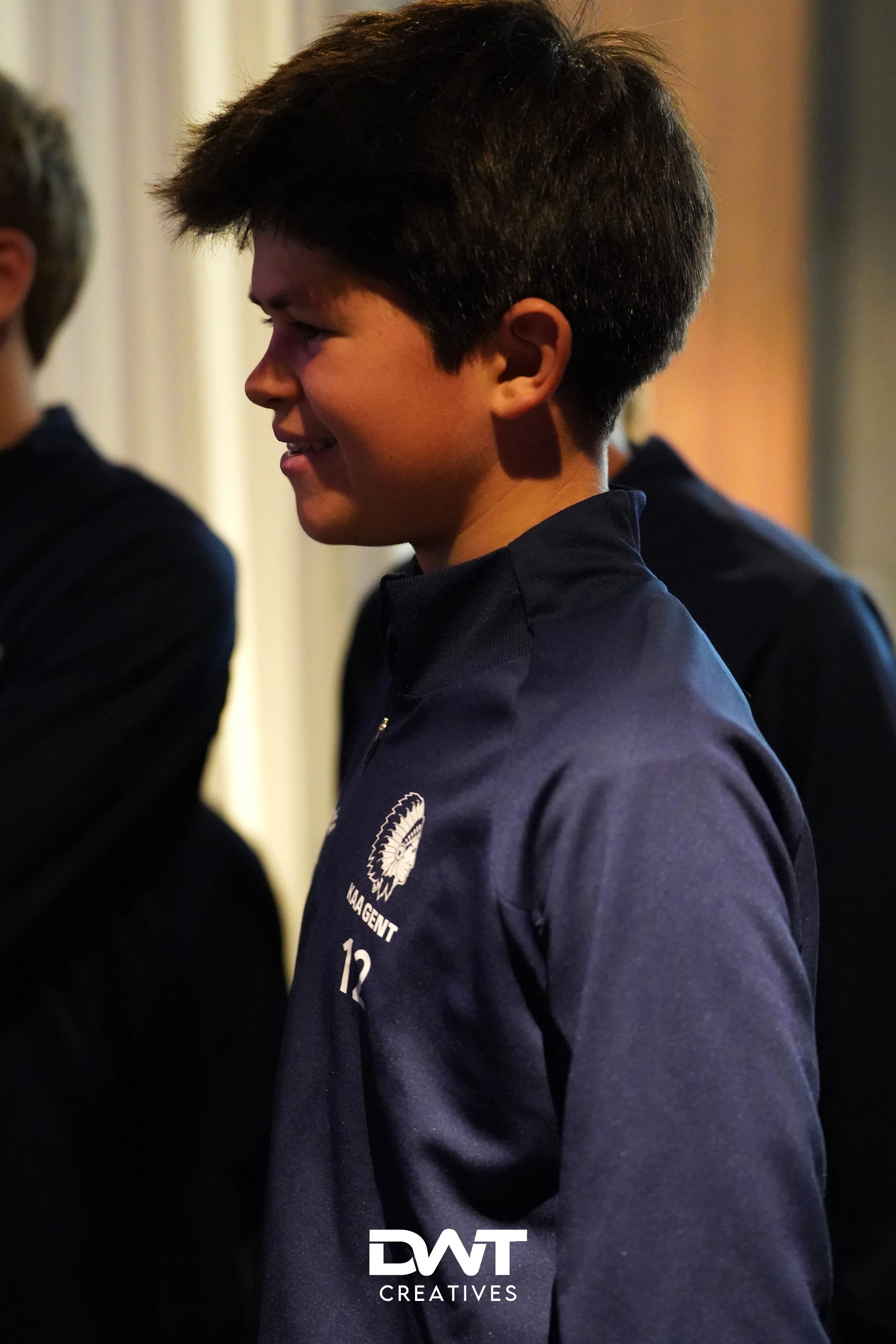 Jongen in donkere trui met logo "KAA Gent" en nummer "12" op de borst, zijkant gefotografeerd.
