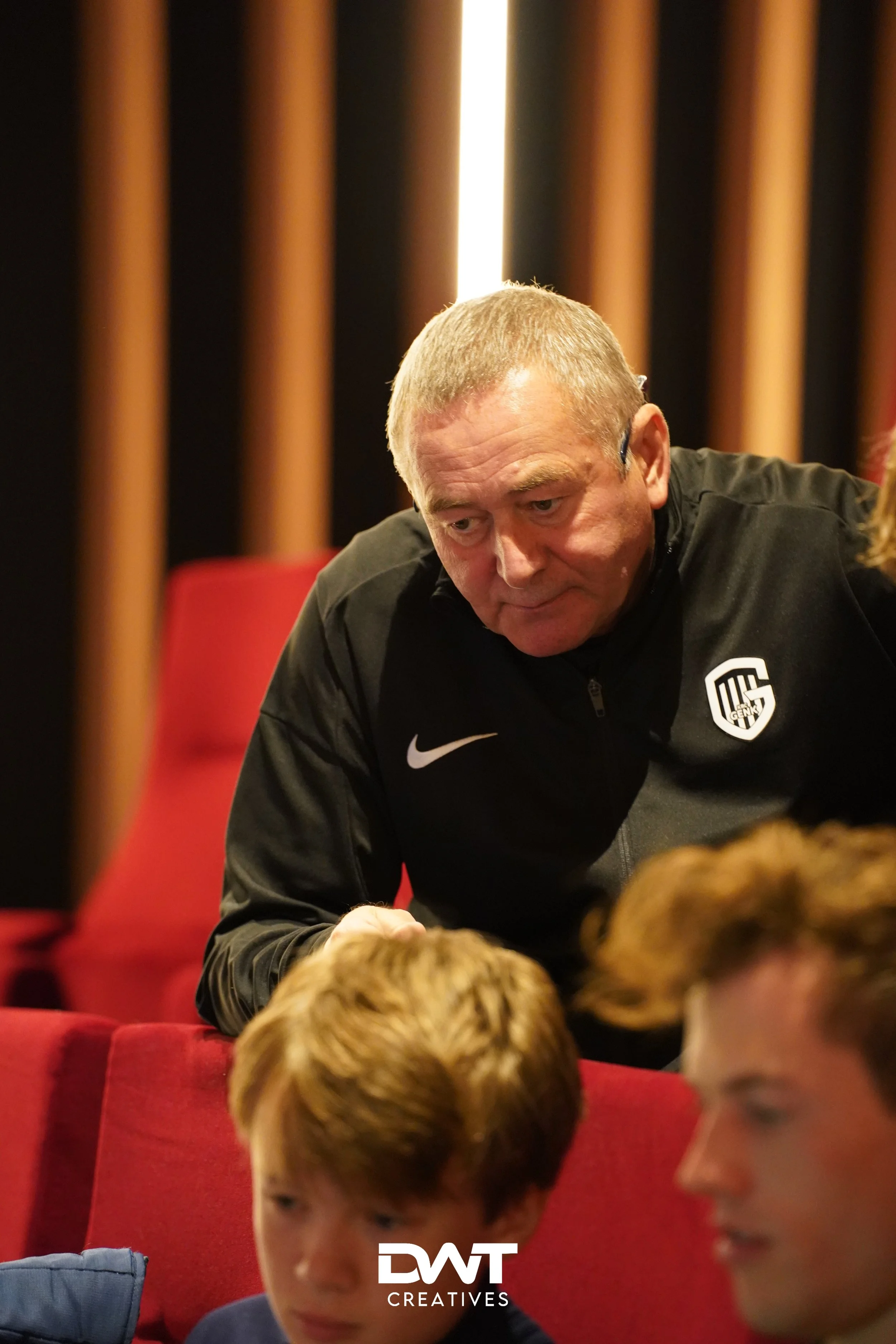 Oudere man in zwarte sportkleding met logo leunt voorover, met twee jongeren op de voorgrond in een omgeving met rode stoelen.