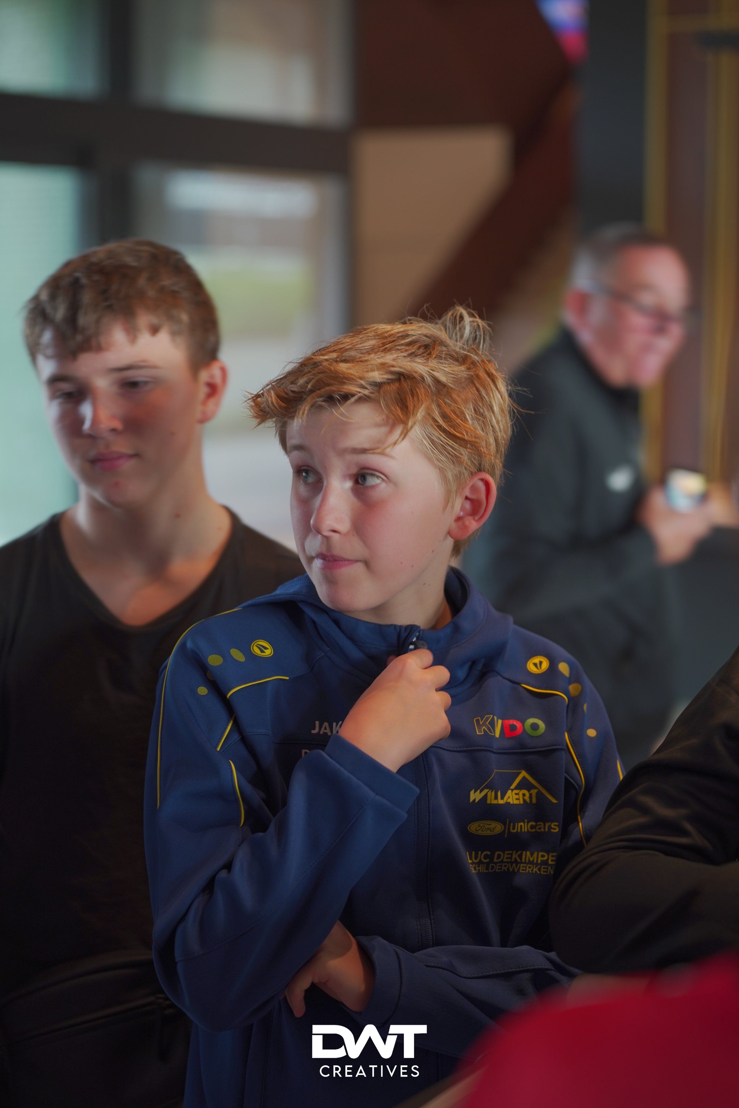 Twee tienerjongens staan binnenshuis, één met een blauwe jas met logo's en een andere persoon is onscherp op de achtergrond.