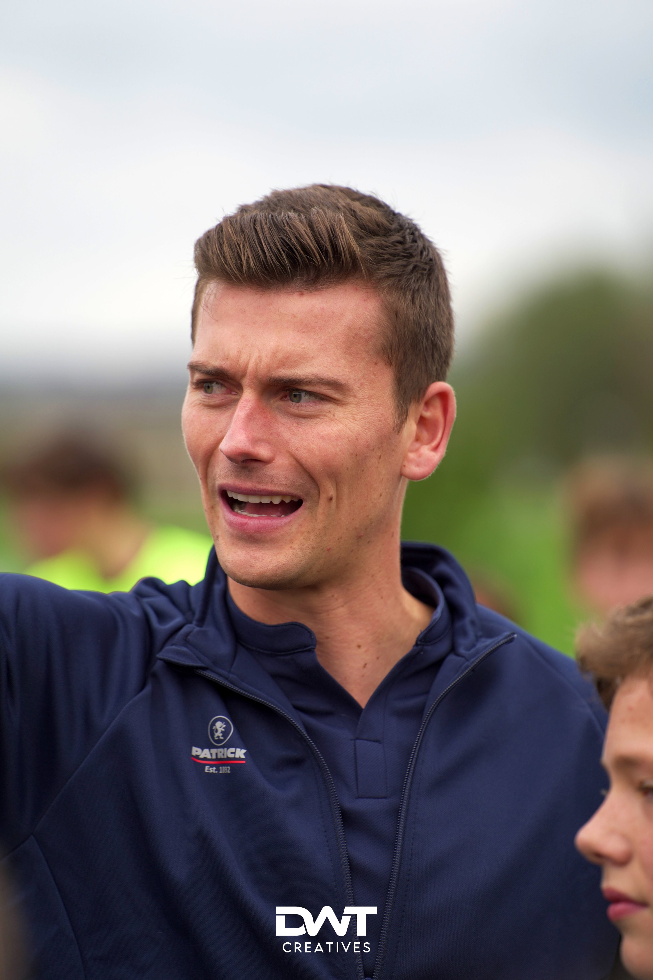 Man in donkerblauwe sportkleding wijst en praat buiten met onscherpe achtergrond.