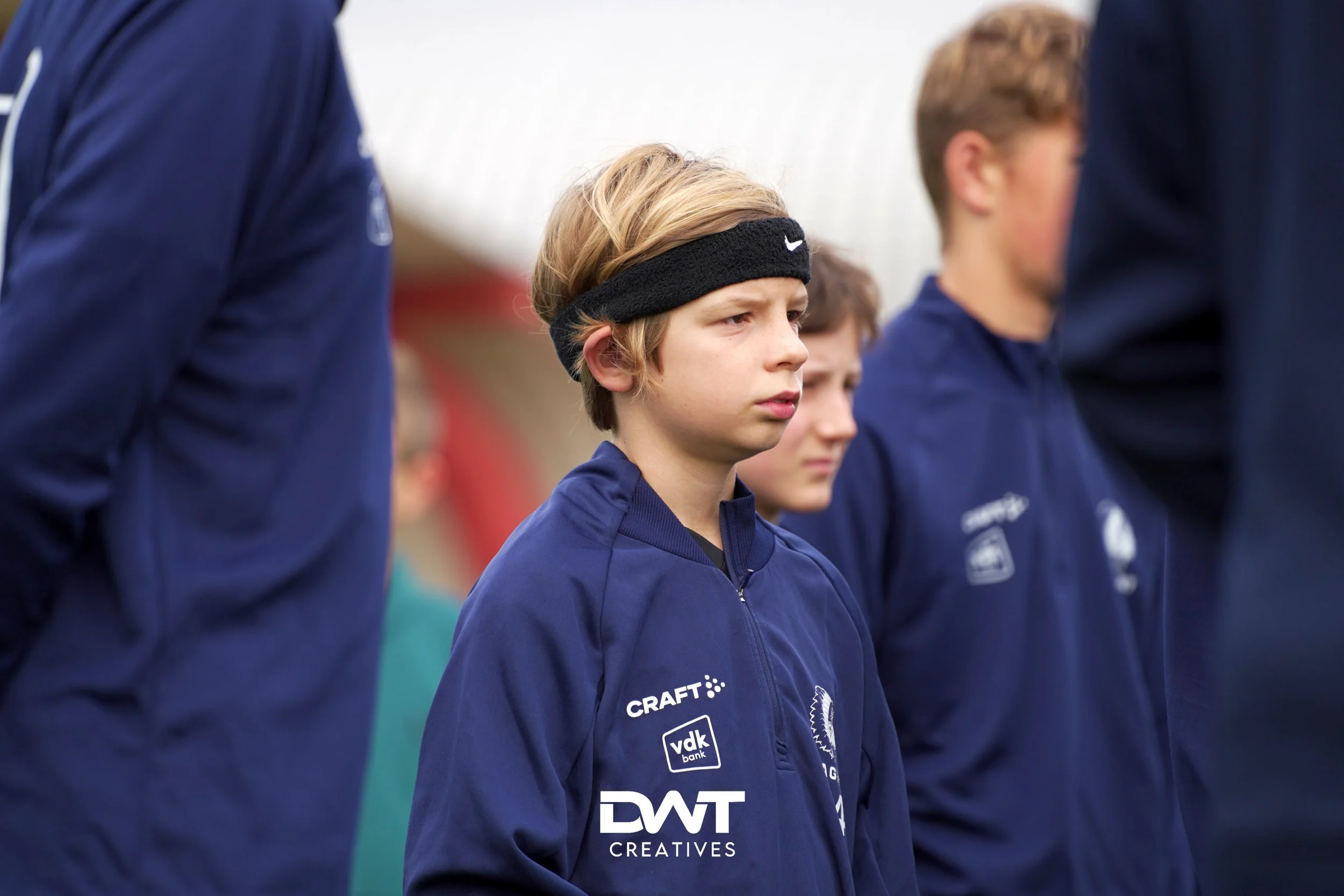 Jongens in sportkleding buiten tijdens een evenement, met een jongen op de voorgrond die een zwart hoofdbandje draagt en een blauw trainingspak met "Craft" en andere logo's.