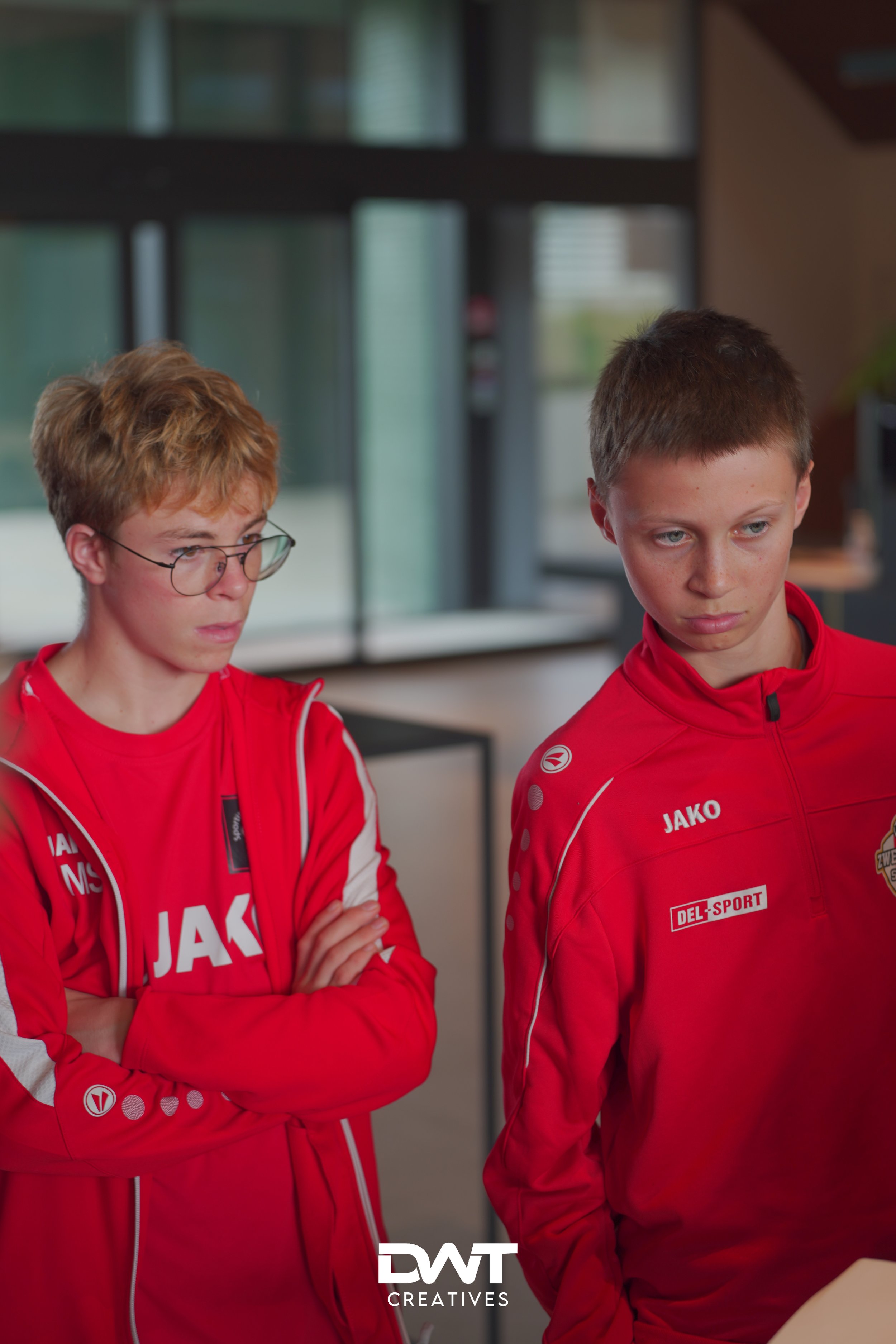 Twee jongeren in rode sportkleding van het merk JAKO staan naast elkaar, mogelijk in een sportieve omgeving of voorbereiding.