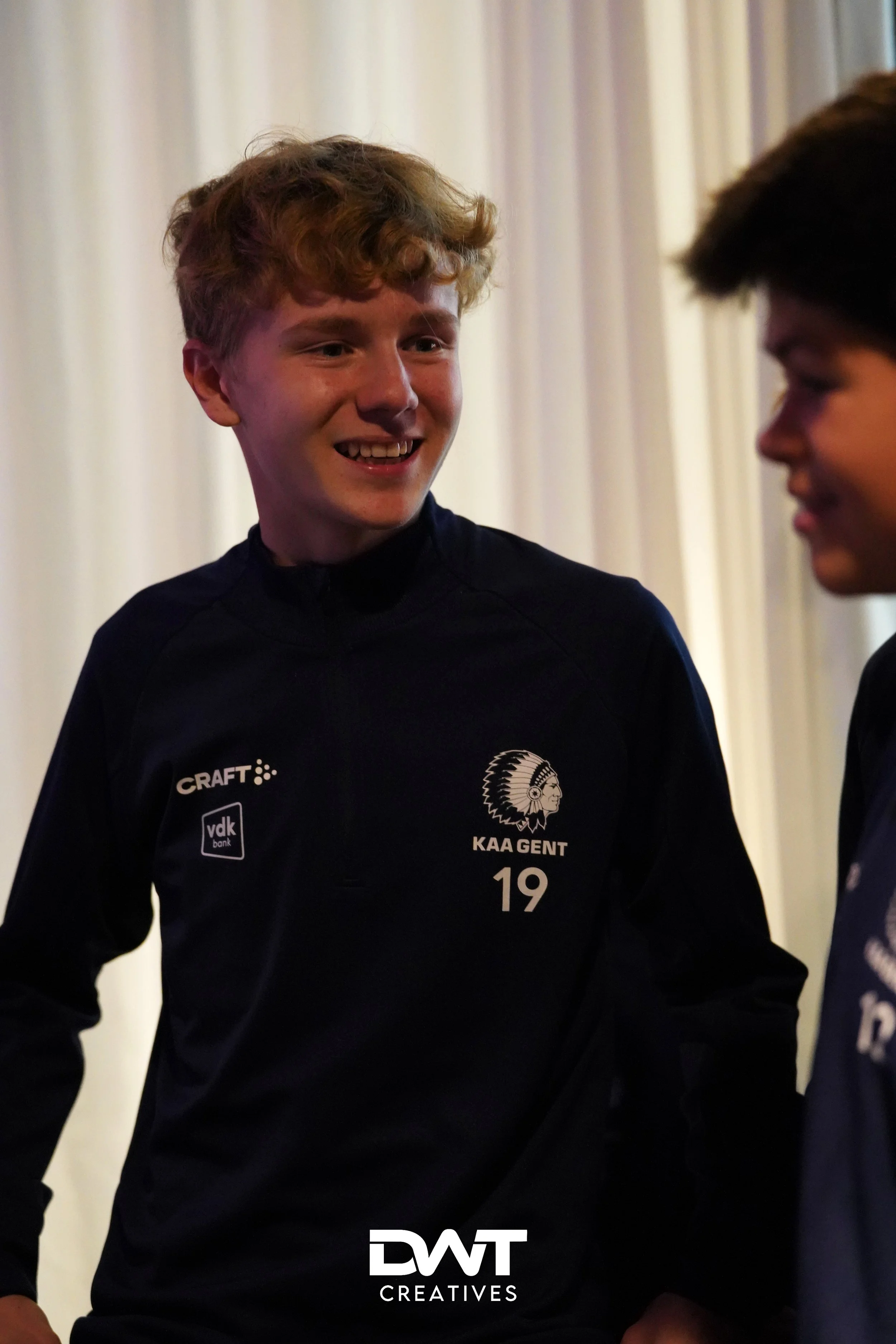 Twee jonge mannen in KAA Gent sportkleding praten en lachen met elkaar, met logo's van Craft en vdk bank zichtbaar.