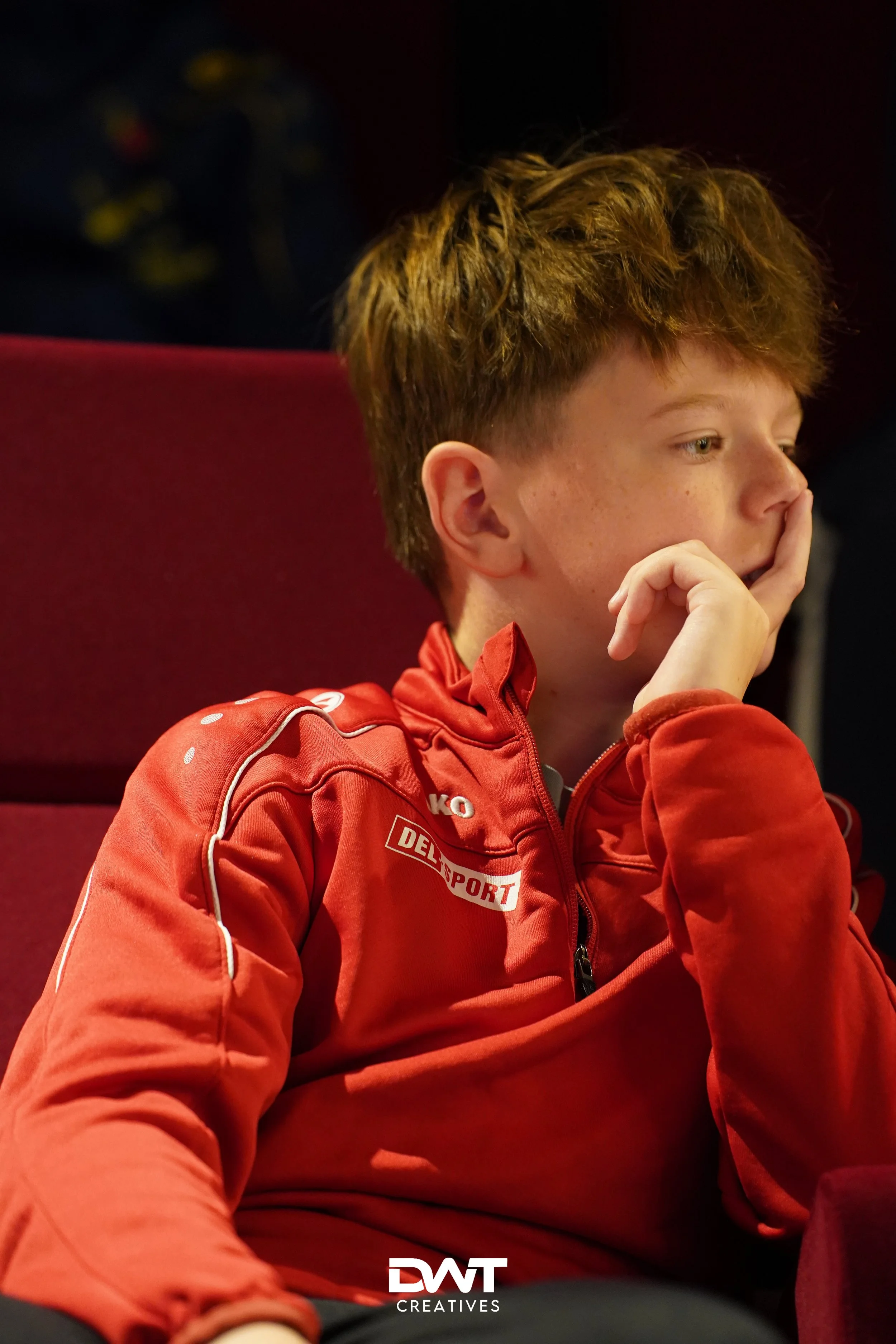 Jongen in rood sportjack, zittend en nadenkend, met hand onder kin, donkere achtergrond.