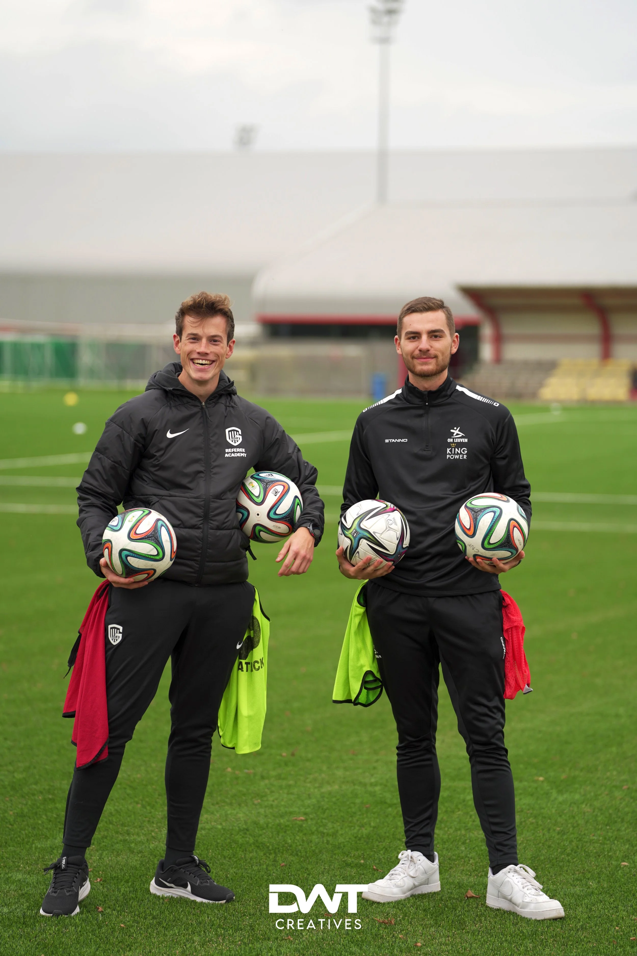 Twee mannen in sportkleding staan op een voetbalveld en houden voetballen vast. Ze dragen trainingsbroeken en jassen met logo's van sportclubs. Achter hen zijn de tribunes van een stadion te zien. Er heerst een ontspannen sfeer.