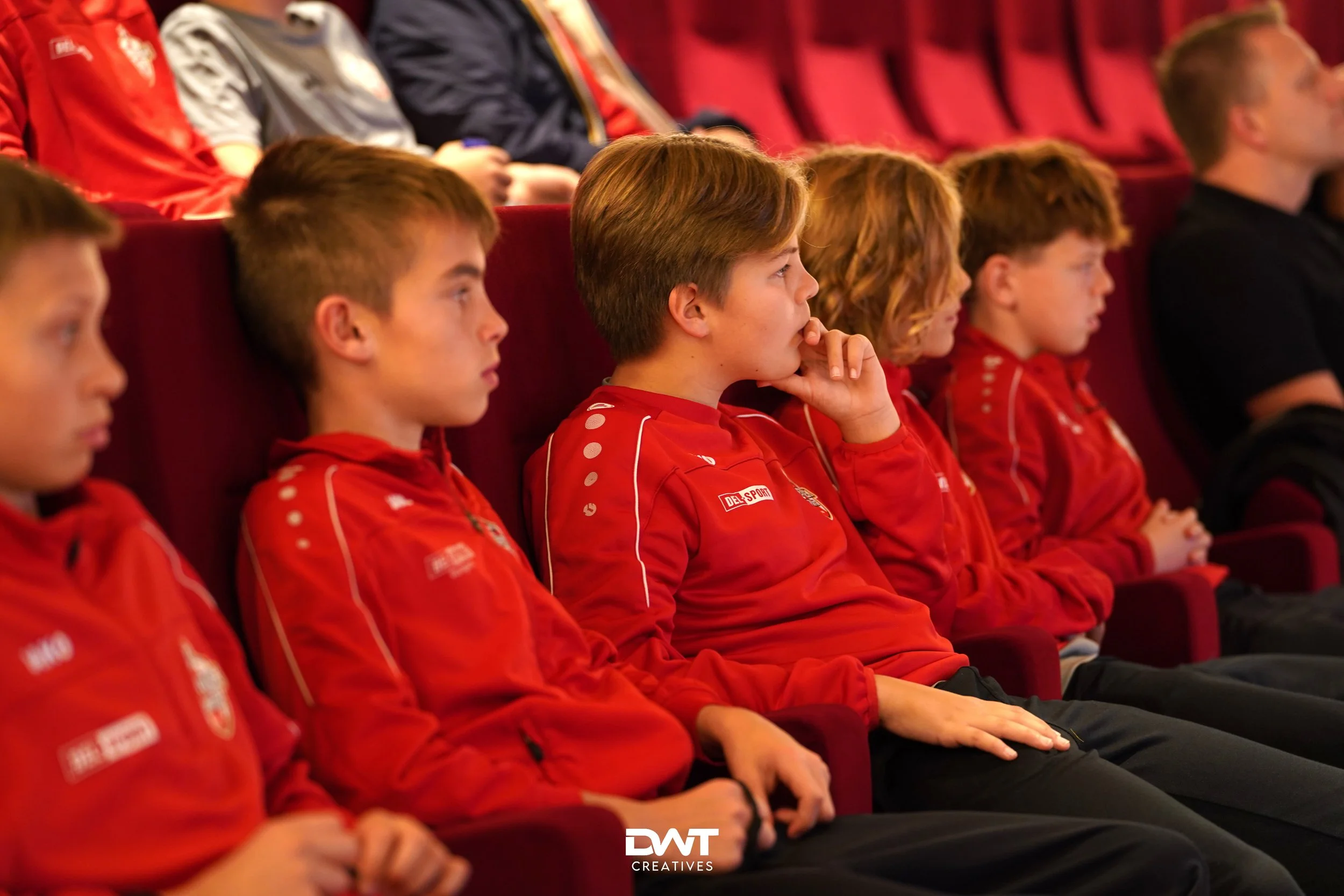 Jonge jongens in rode sportkleding zitten in een theaterzaal en luisteren aandachtig.