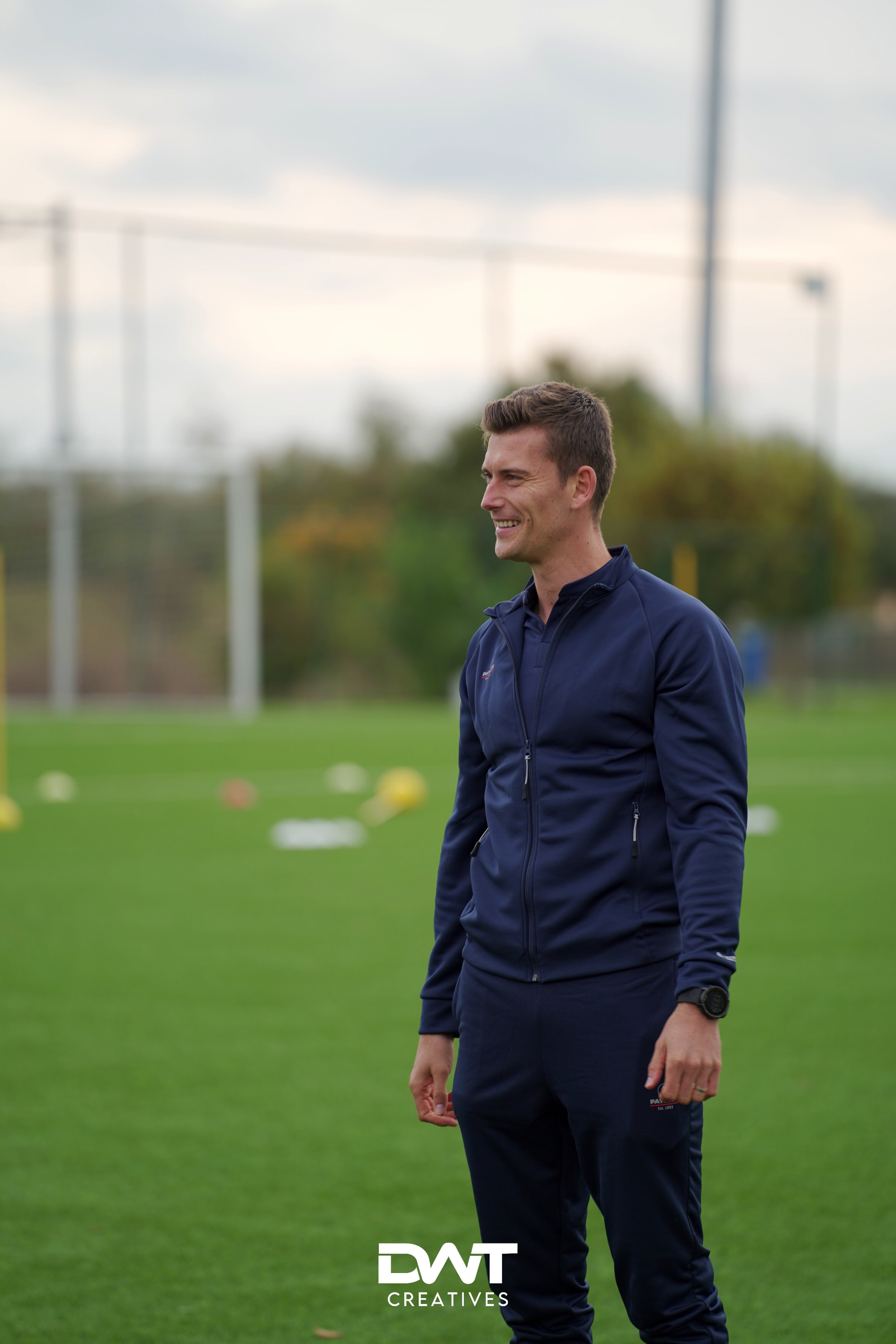 Man in sportkleding op een voetbalveld, lachend met groene grasmat op de achtergrond en doelpalen.