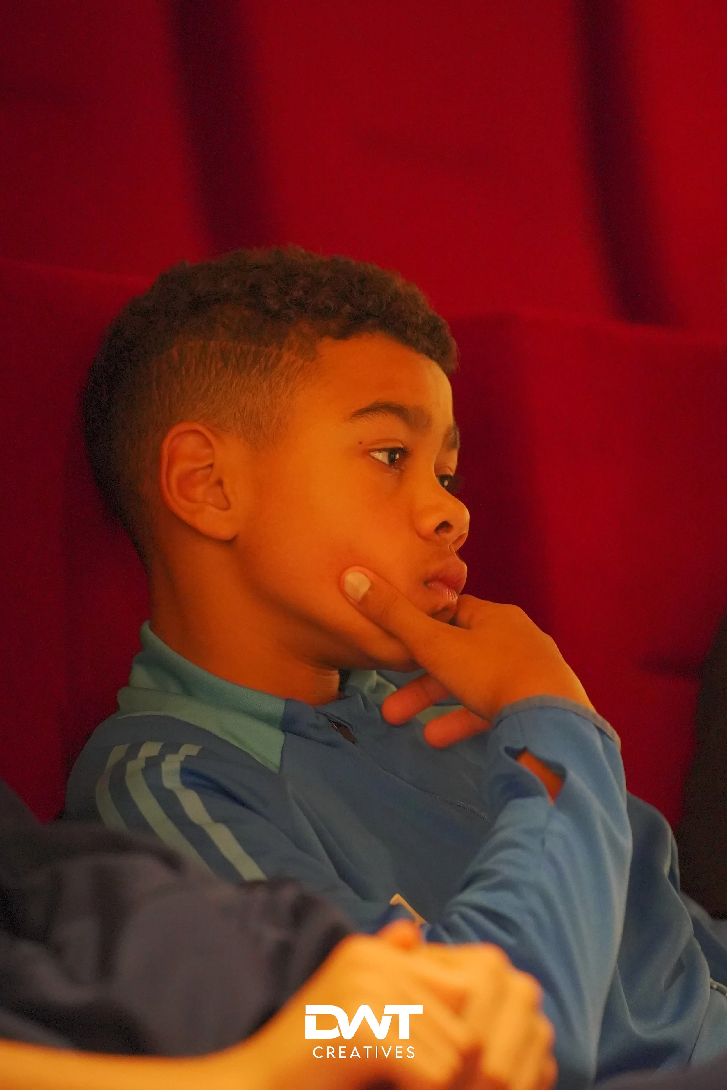 Jongen in blauwe kleding kijkt geconcentreerd naar iets, met zijn hand op zijn kin tegen een rode achtergrond.