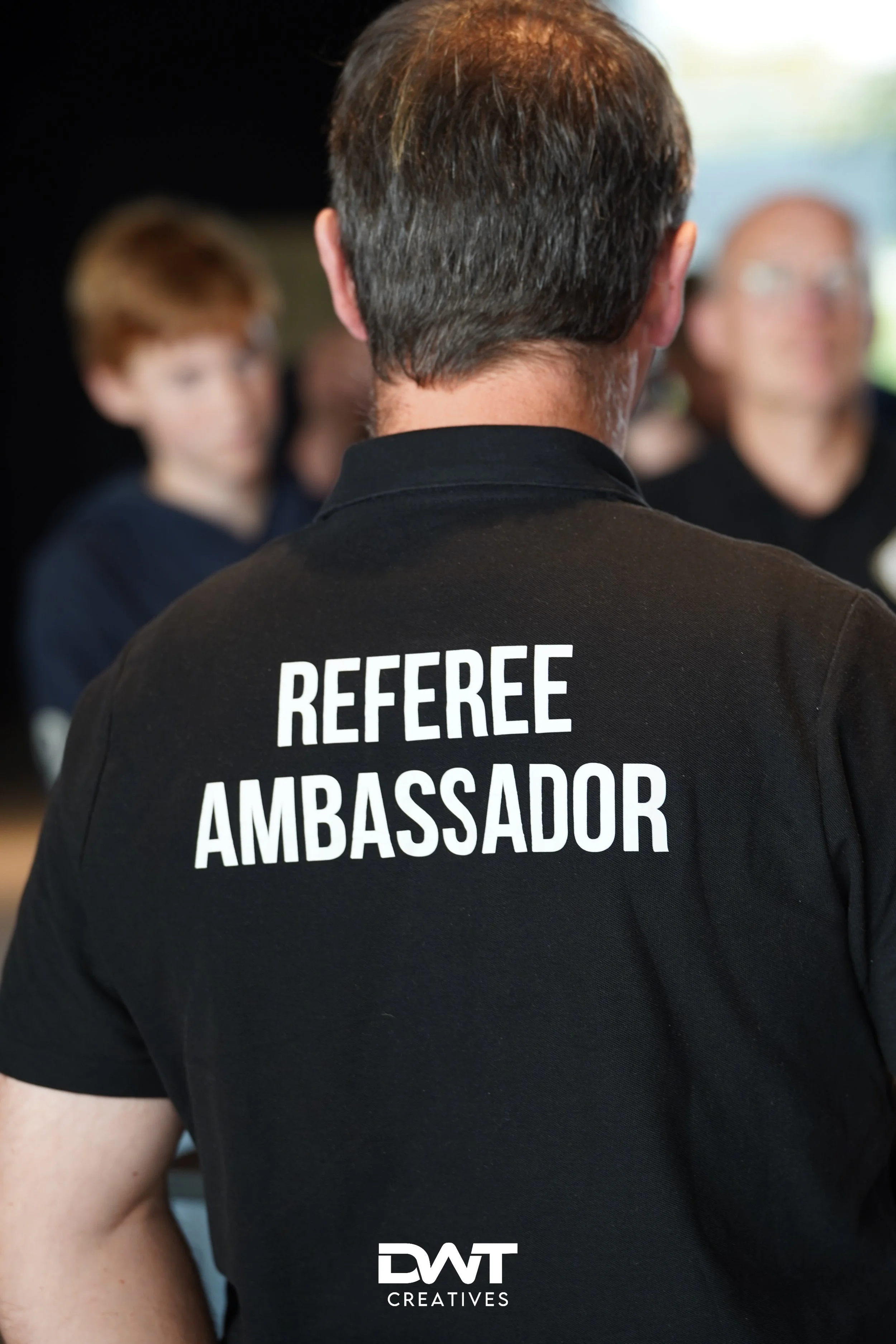 Man met zwart shirt met 'Referee Ambassador' erop, gefotografeerd van achteren in een groepssetting.