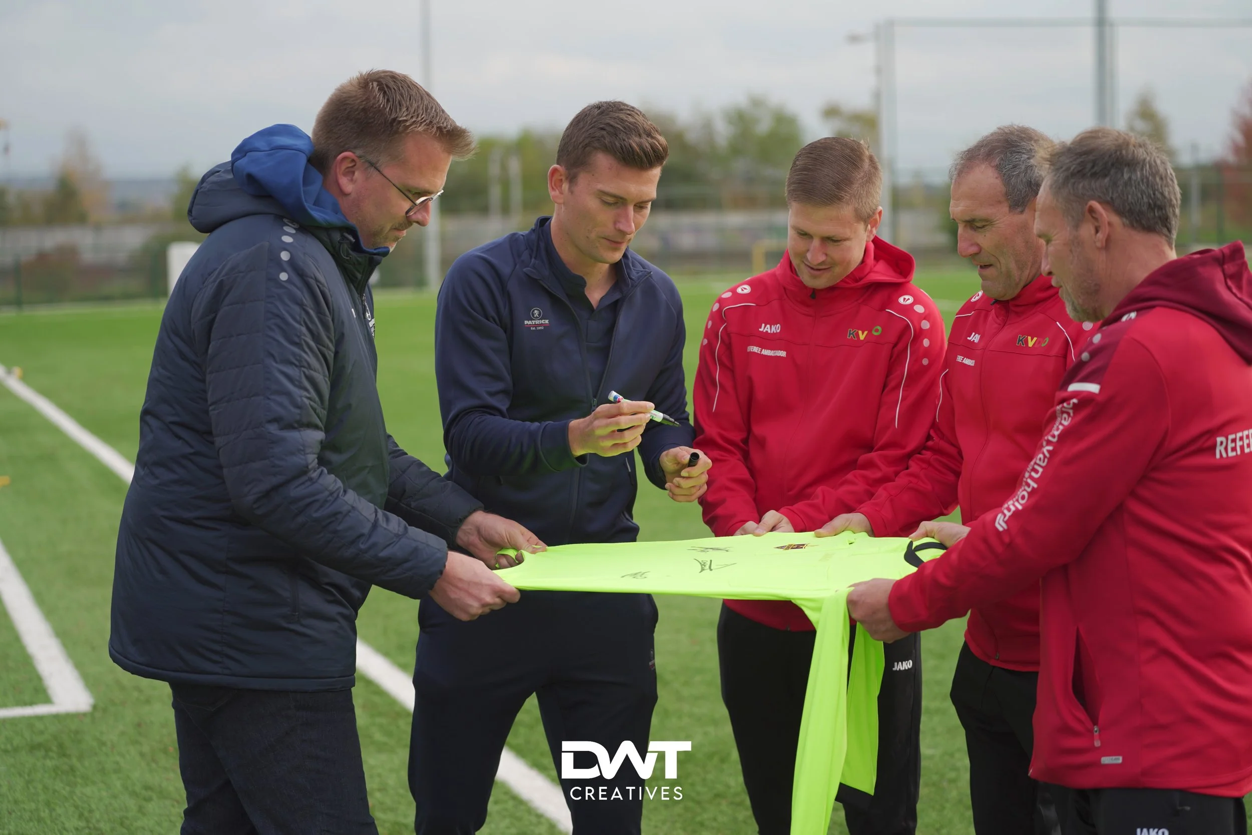 Mensen staan op een voetbalveld en bekijken een felgekleurd sportshirt dat wordt vastgehouden voor een handtekening. Ze dragen sportkleding en lijken betrokken bij een sportevenement.
