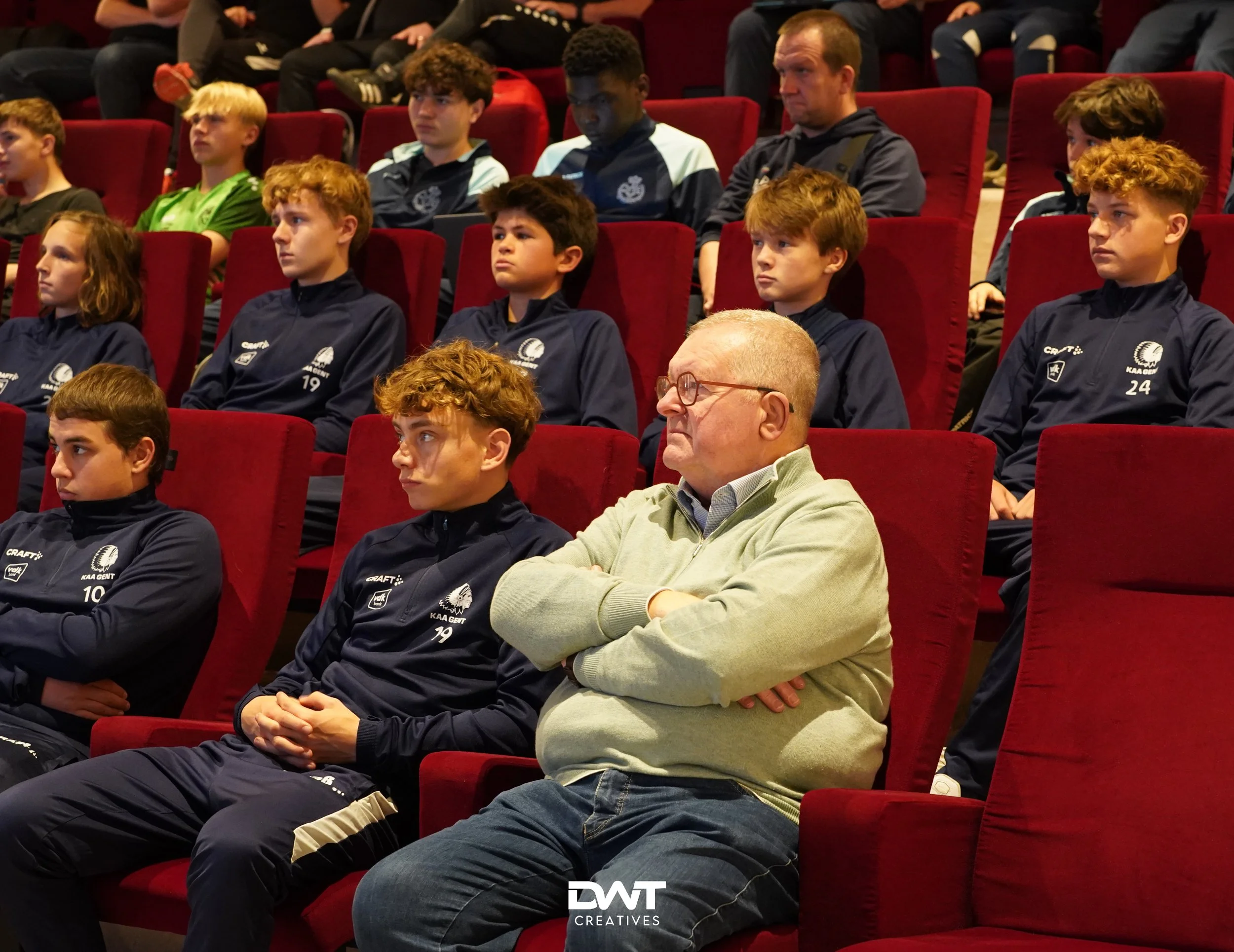 Mensen zitten in rode stoelen tijdens een evenement, met jonge mannen in sportkleding en een oudere man in een trui. Ze kijken geconcentreerd naar een presentatie.