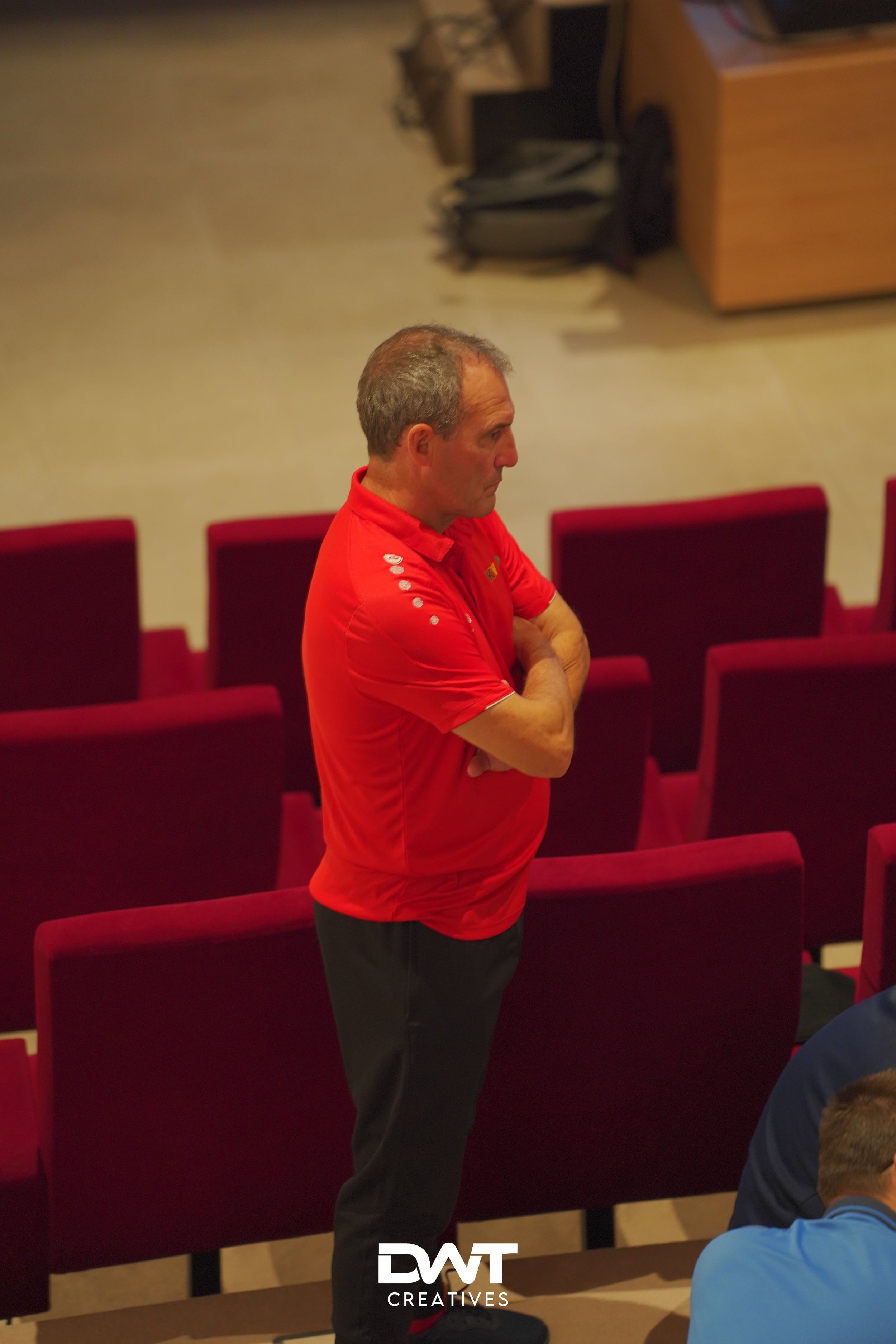 Man in rood shirt staat met armen over elkaar in zaal met rode stoelen.