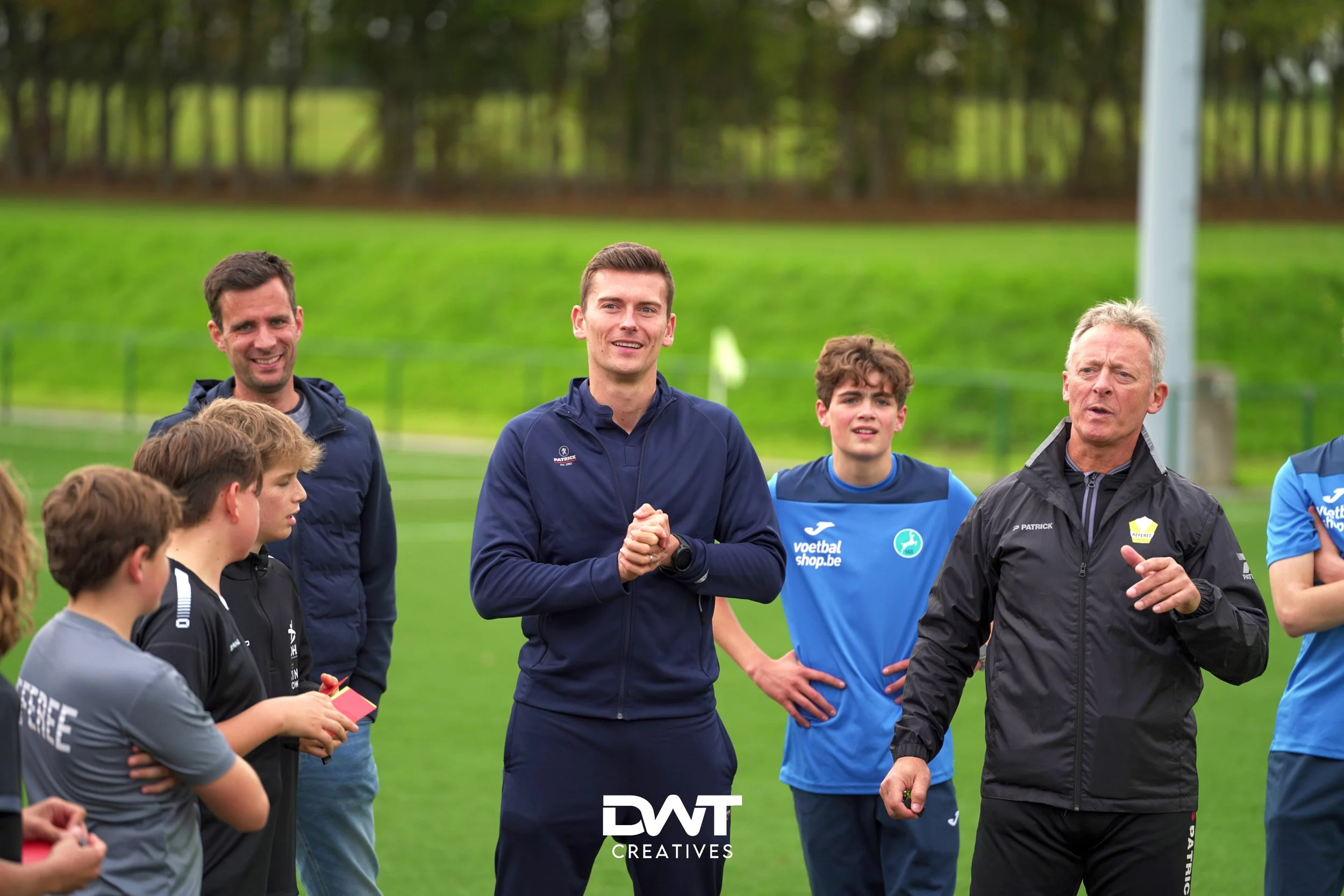 Een groepje mensen op een voetbalveld tijdens een trainingssessie. Een man spreekt tot de groep. Enkele jongeren luisteren aandachtig. DWT Creatives logo onderaan.