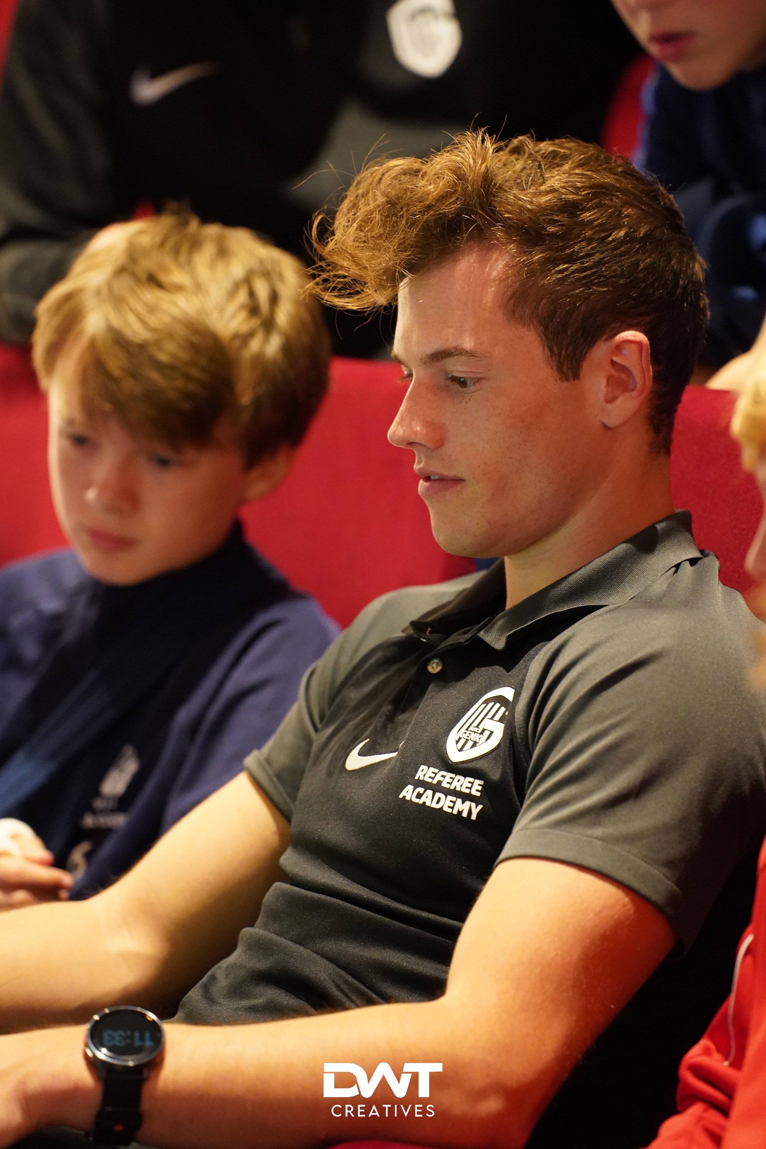 Groep mensen in een binnenruimte, gefocust op een jongeman met een "Referee Academy" shirt. Er zijn kinderen bij hem in de buurt.