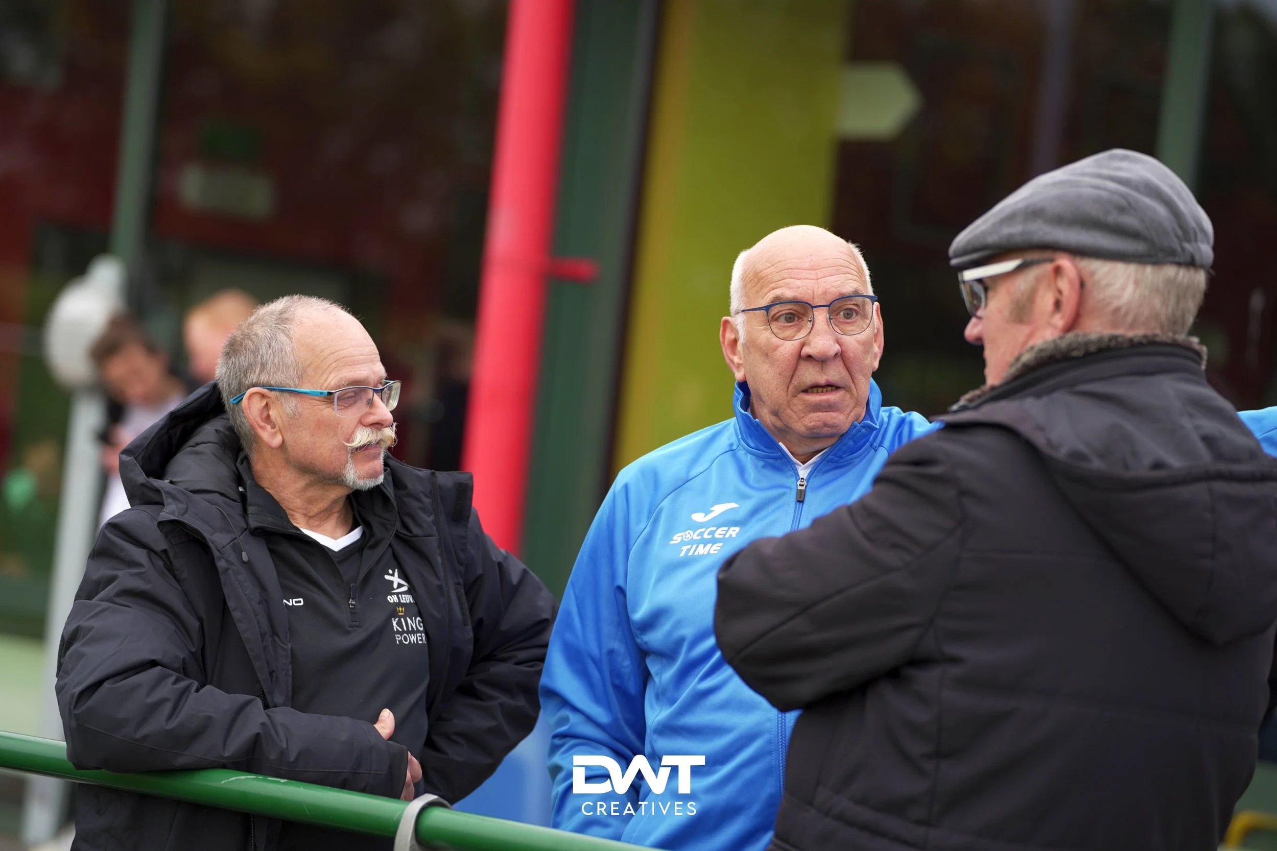 Drie oudere mannen in gesprek buiten, twee van hen dragen sportkleding en de derde draagt een jas en pet. Op de achtergrond is een gebouw zichtbaar.
