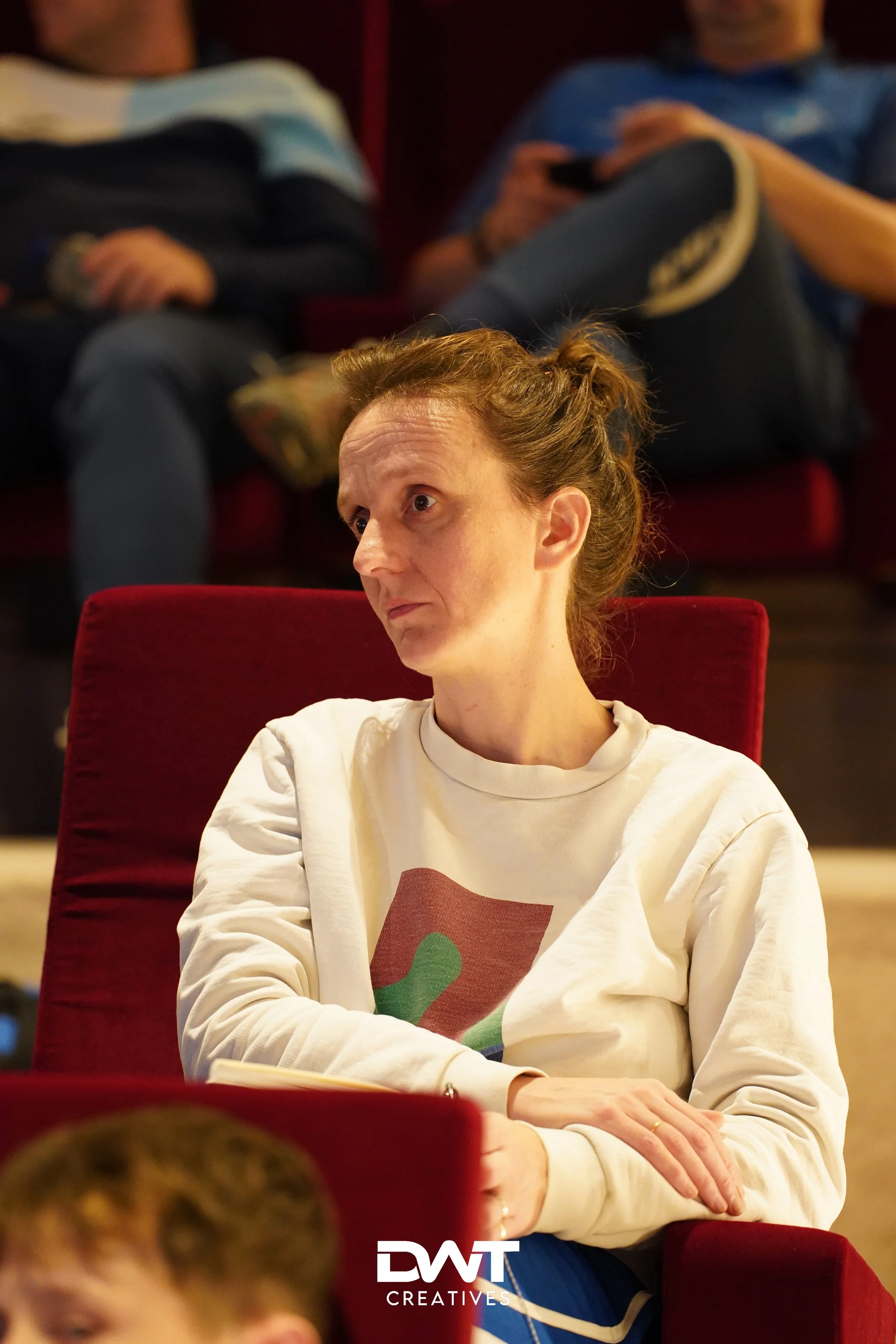 Vrouw in witte trui zit op rode stoel, kijkt aandachtig richting voren. Personen op achtergrond zitten ontspannen. Geheel binnenomgeving.