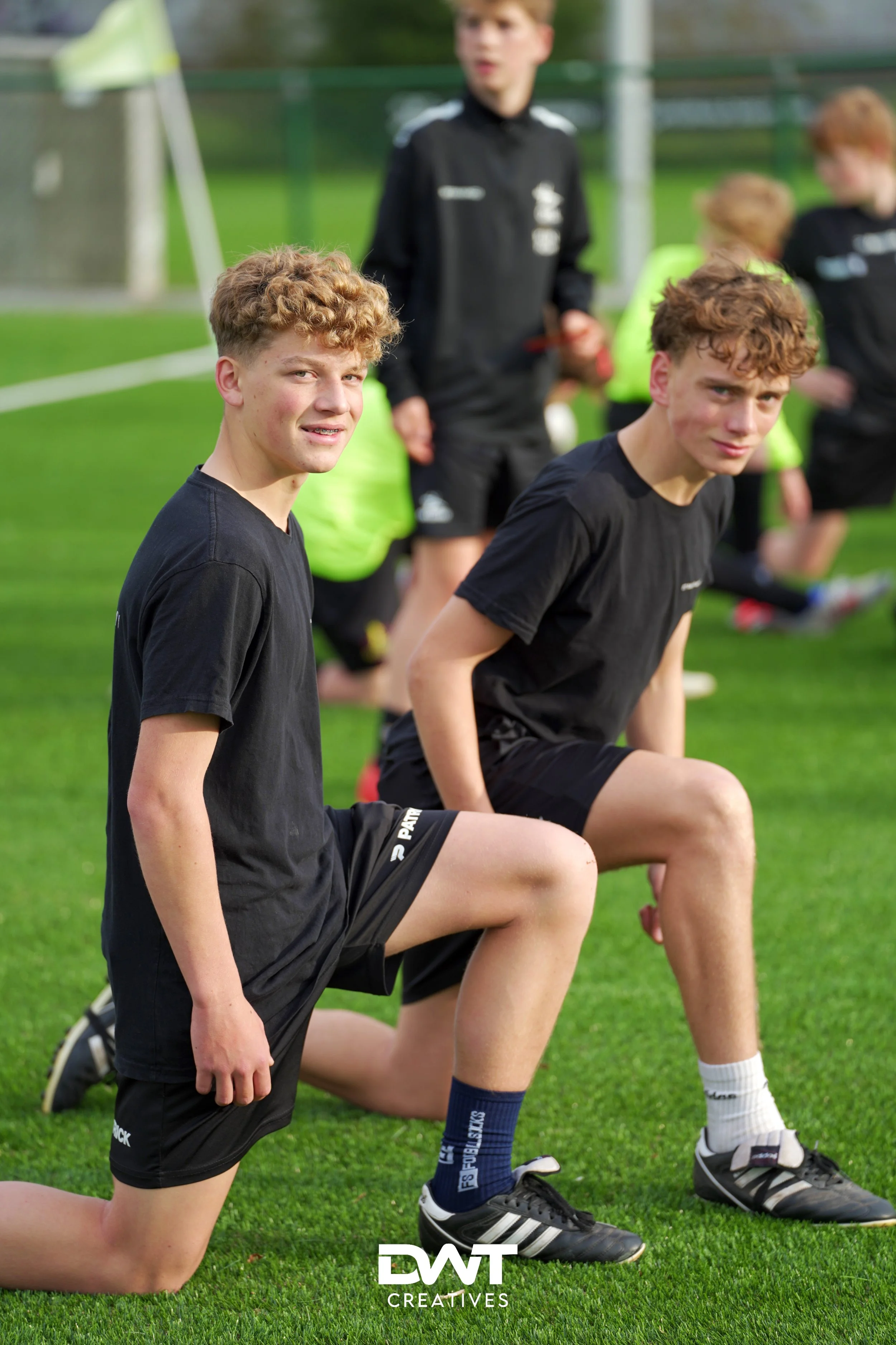 Jonge voetballers tijdens een training op een grasveld, gekleed in sportkleding en knielend terwijl ze naar de camera kijken.