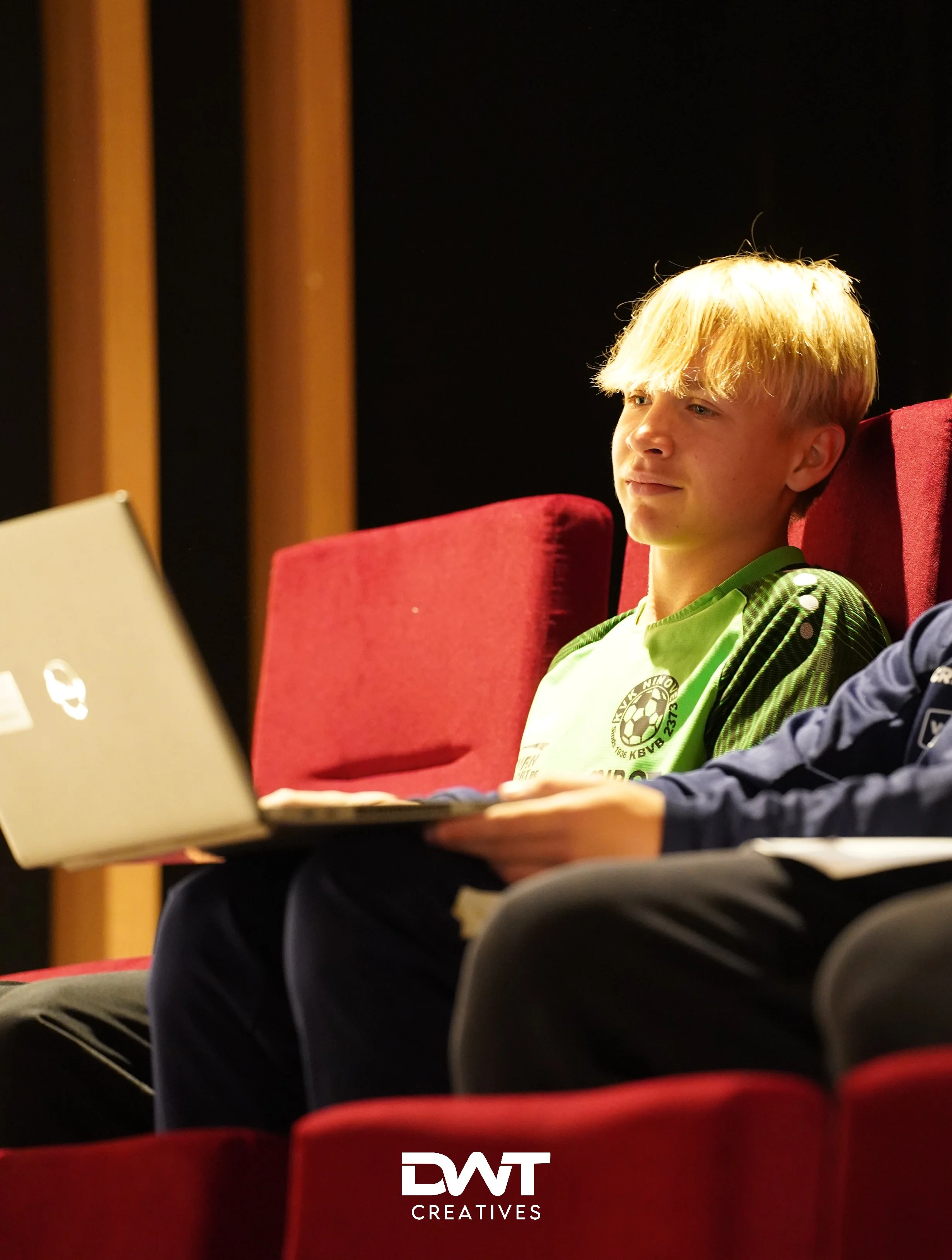 Jongen in groene sportkleding, zittend in rode stoel met laptop, indoor omgeving.