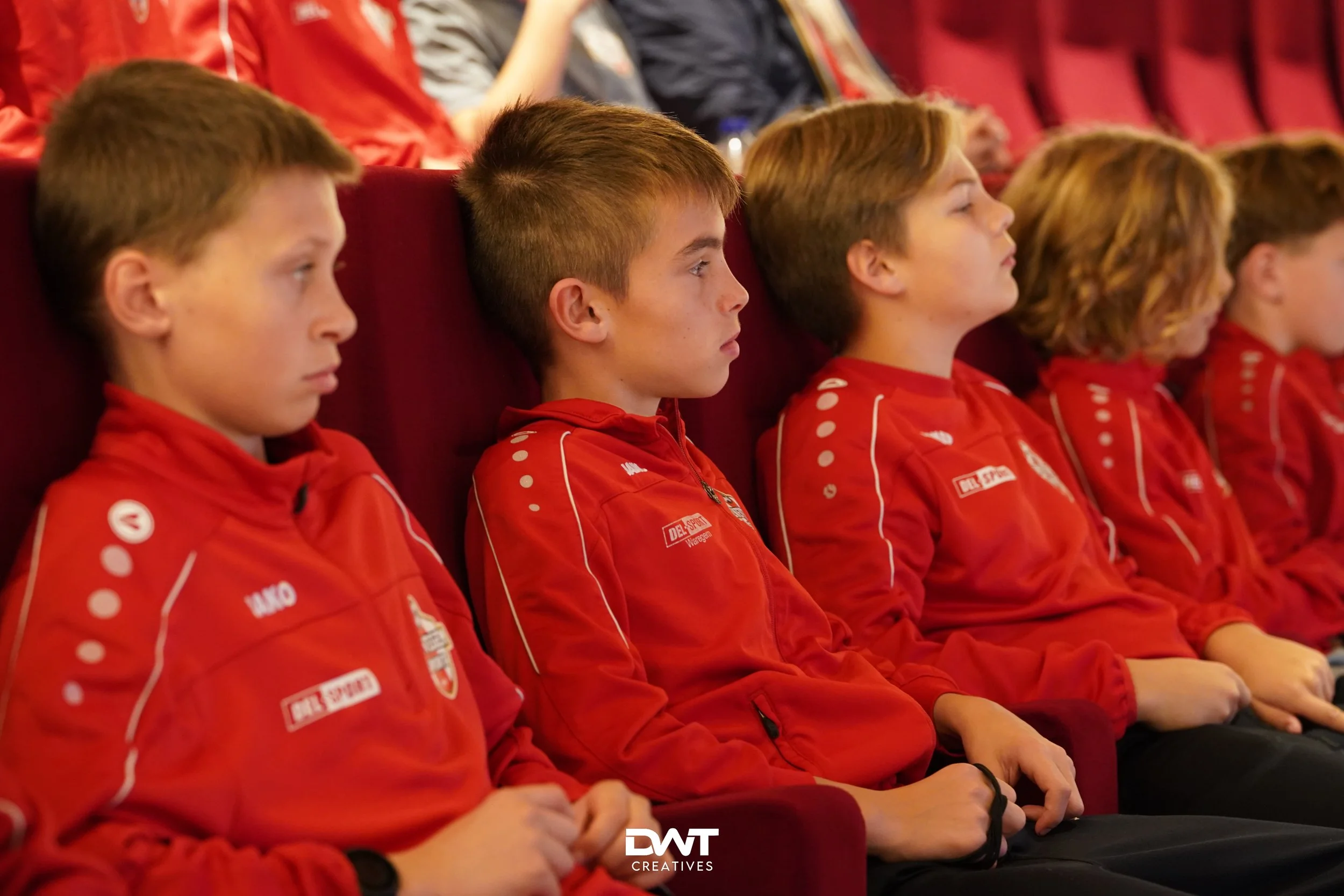 Groep jonge jongens in rode sportkleding zit aandachtig in een auditorium.