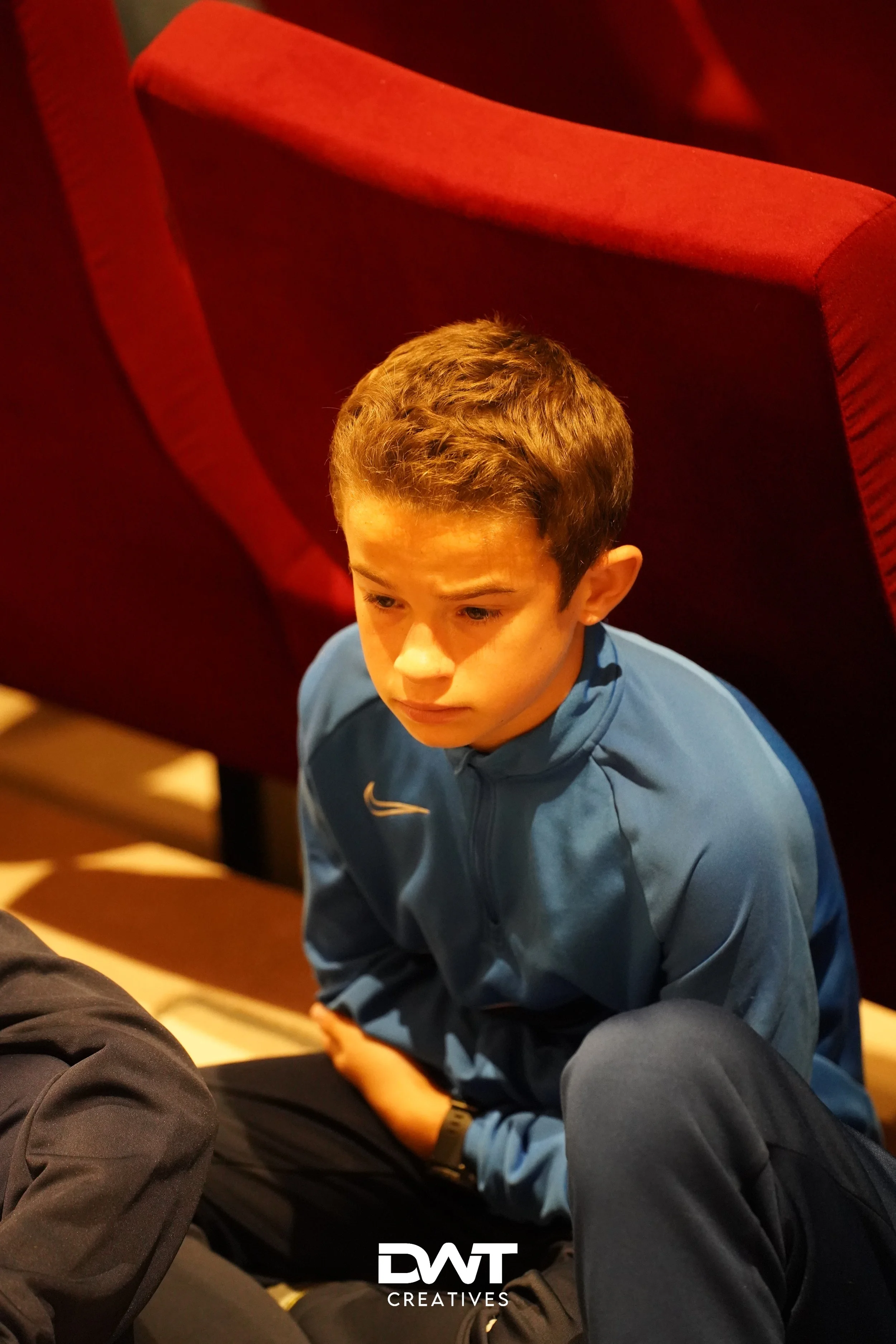 Een jonge jongen in een blauwe sportjas zit naast een rode stoel en kijkt naar beneden, ergens in een indoor omgeving zoals een theater of conferentiezaal.