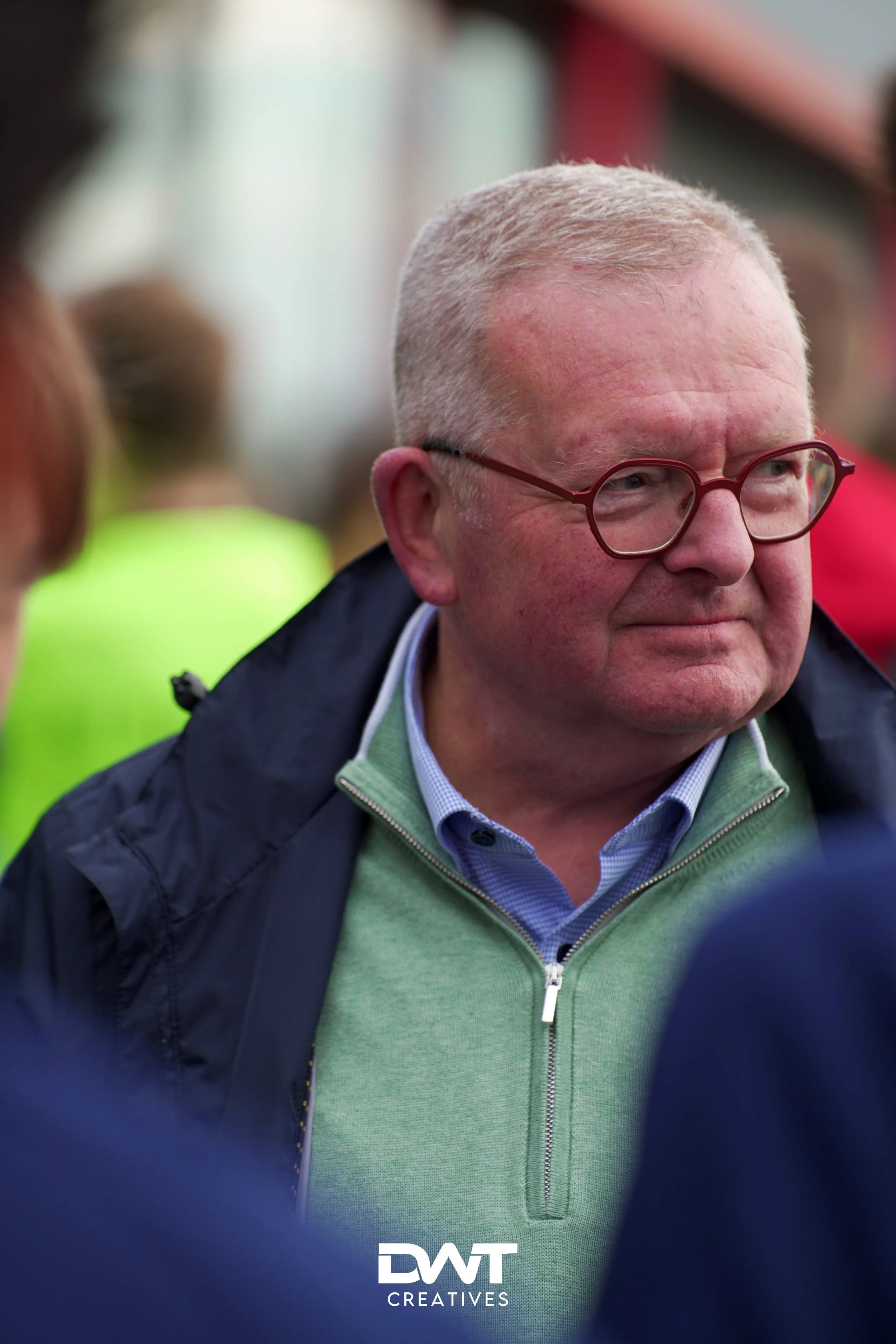 Een oudere man met een bril en een groene trui, in een menigte.