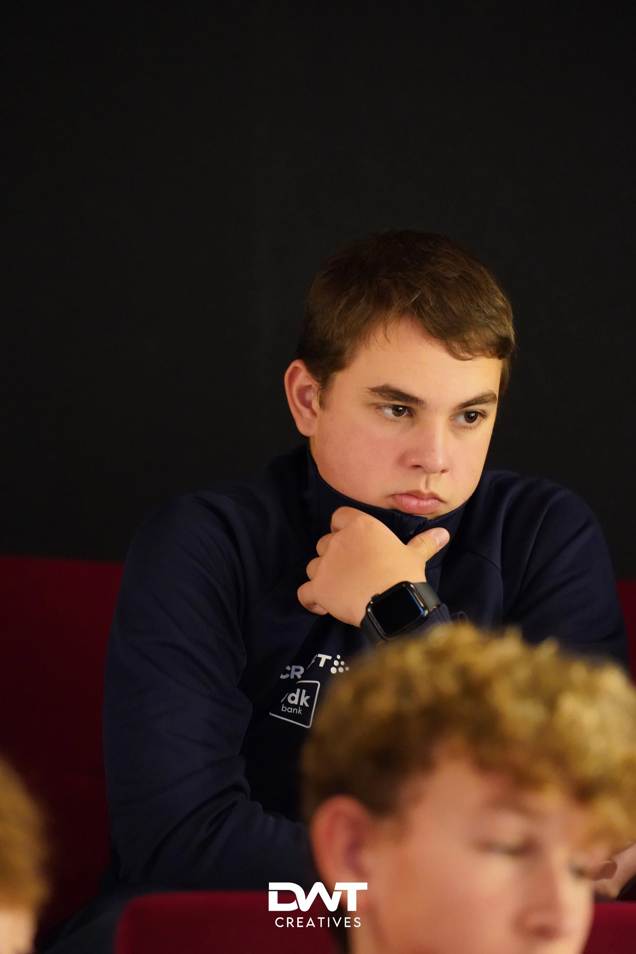 Jongeman in donkerblauwe trui kijkt nadenkend, met hand bij kin, op de achtergrond is een persoon onscherp. Logo DWT Creatives onderaan.