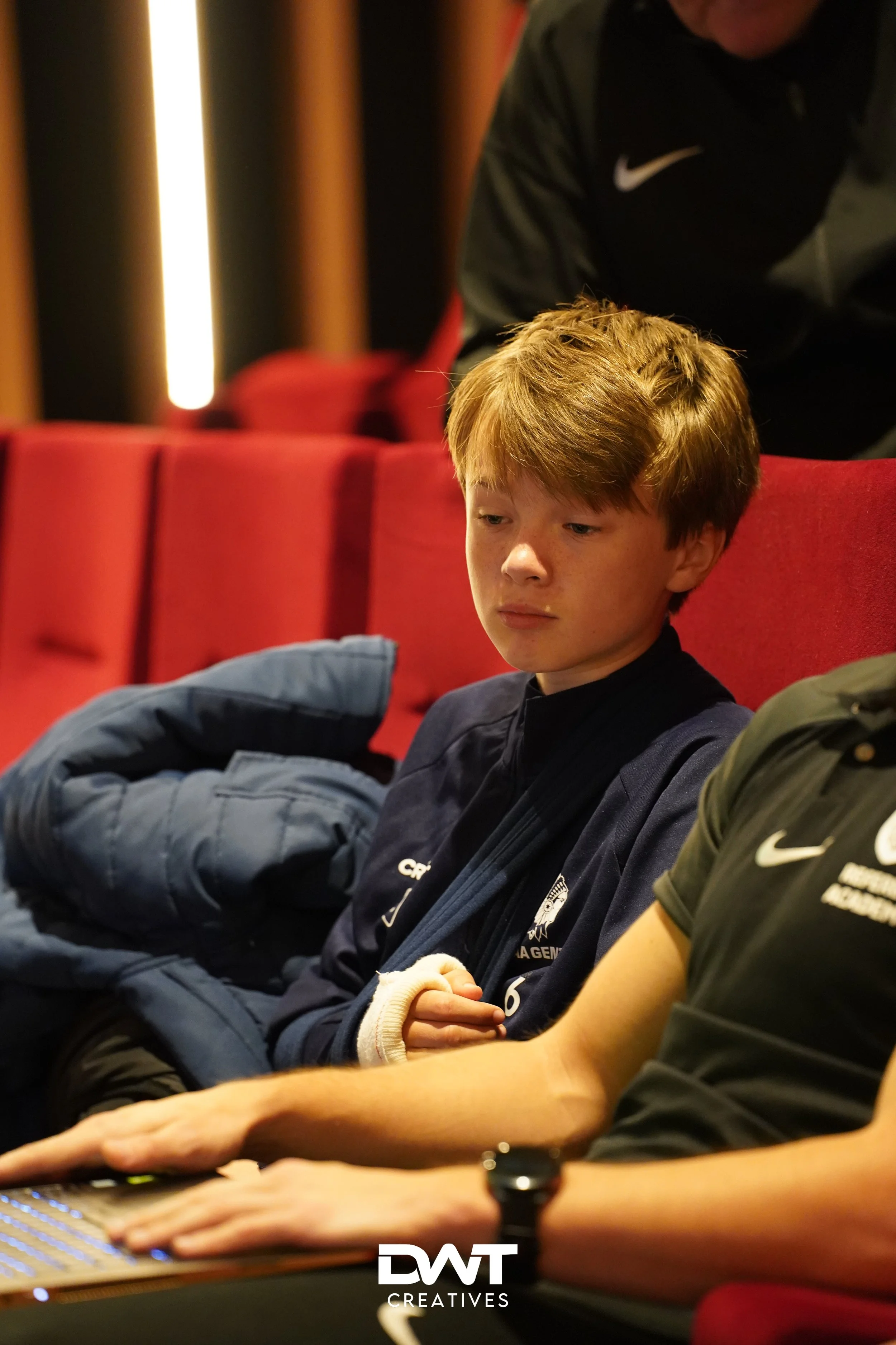 Jongen zit op rode bioscoopstoel, met blauwe jas en verband om hand. Persoon naast hem werkt op laptop. Nike-logo aanwezig.