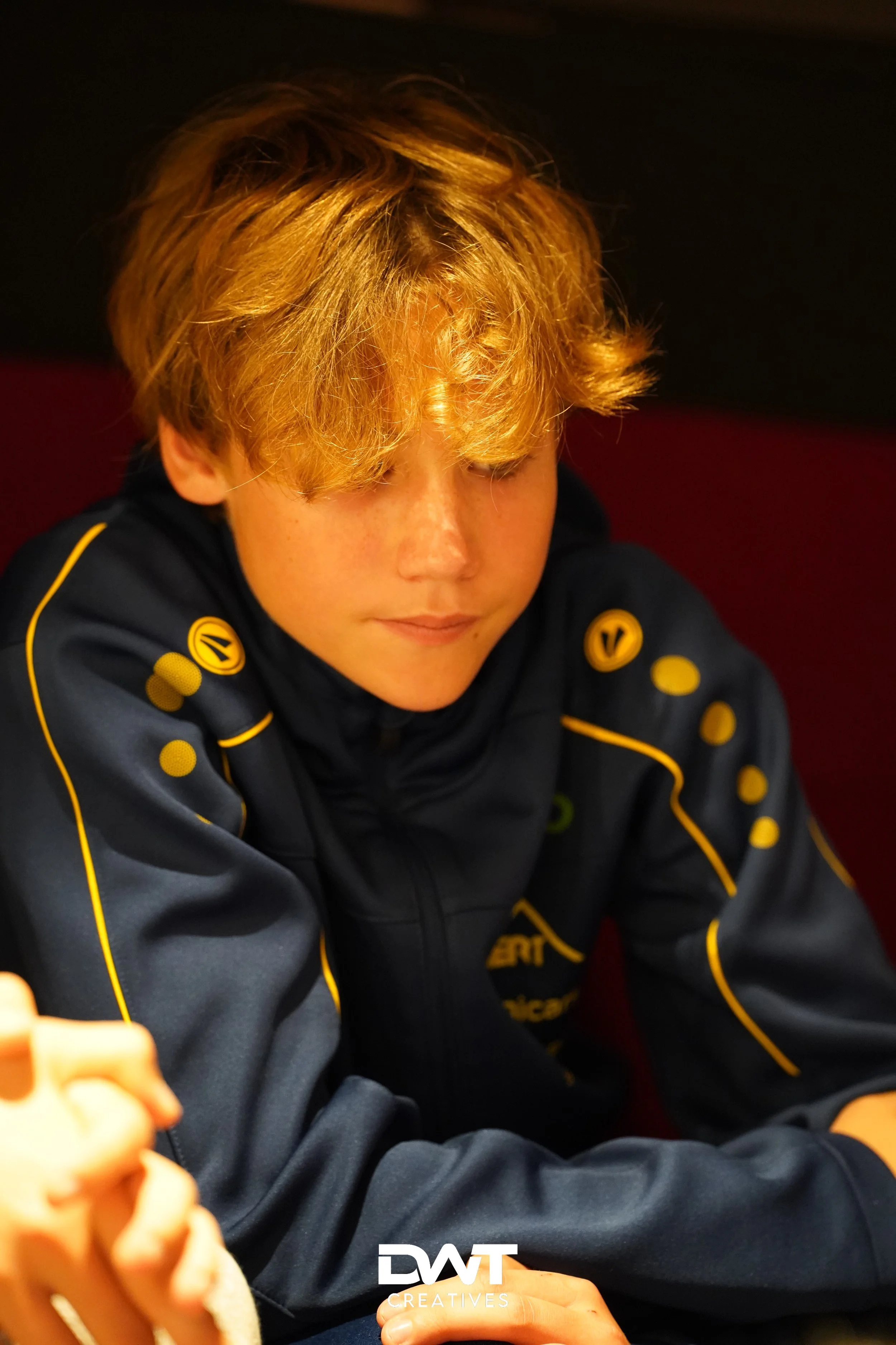 Jongen met halflang blond haar in een blauw sportjack met gele accenten, binnenomgeving.