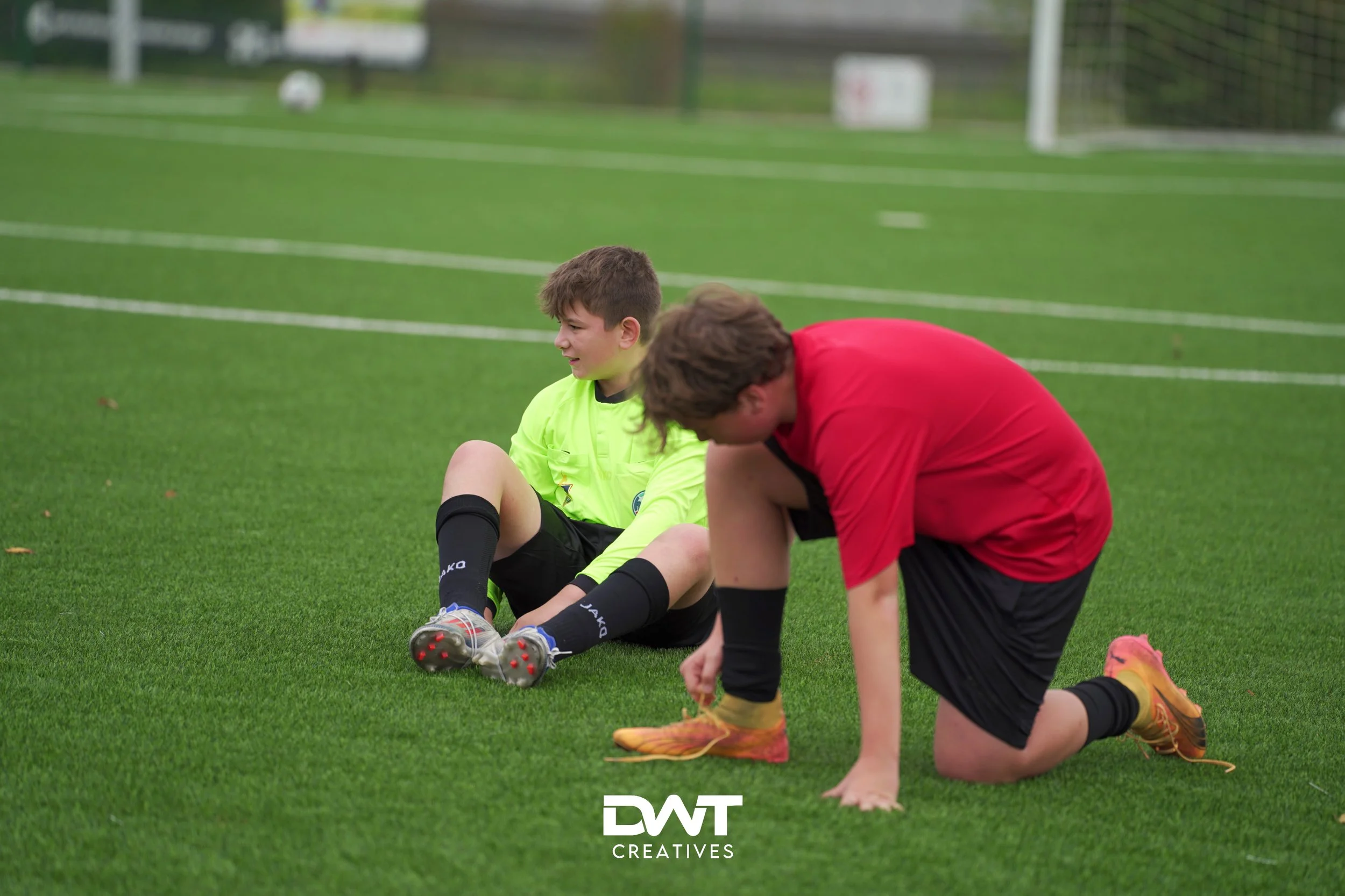 Twee jongens op een voetbalveld; een zit op de grond in een groen shirt, de ander knielt in een rood shirt.