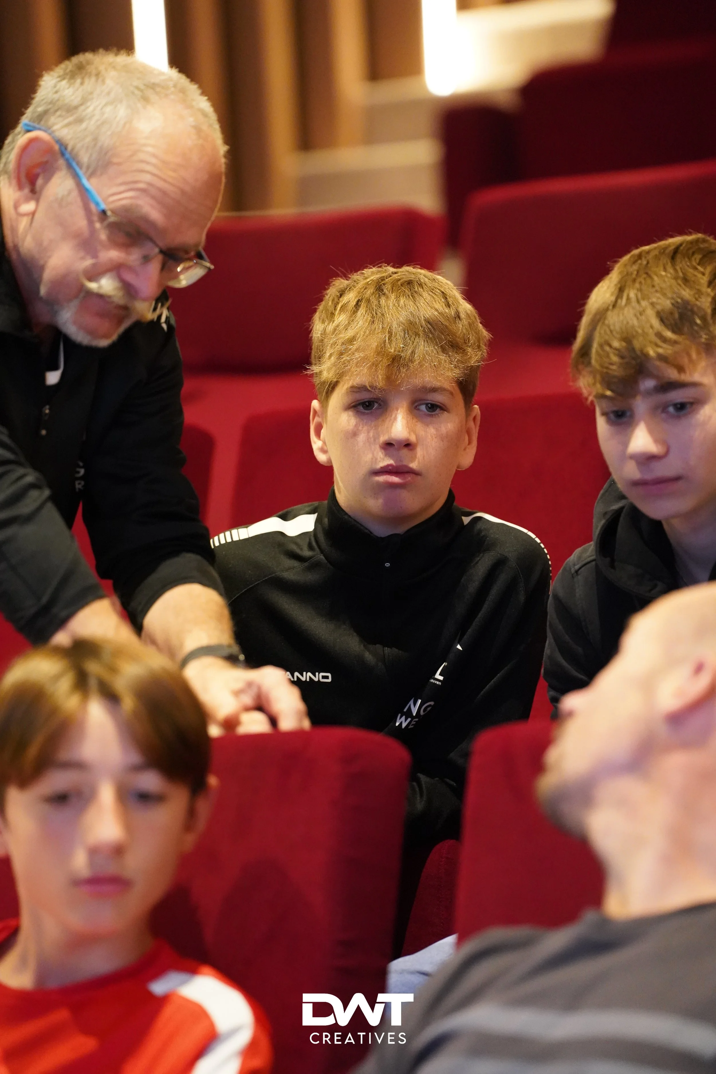 Groep mensen in een theaterzaal met rode stoelen, waaronder een oudere man met bril en vier jongeren die aandachtig kijken.