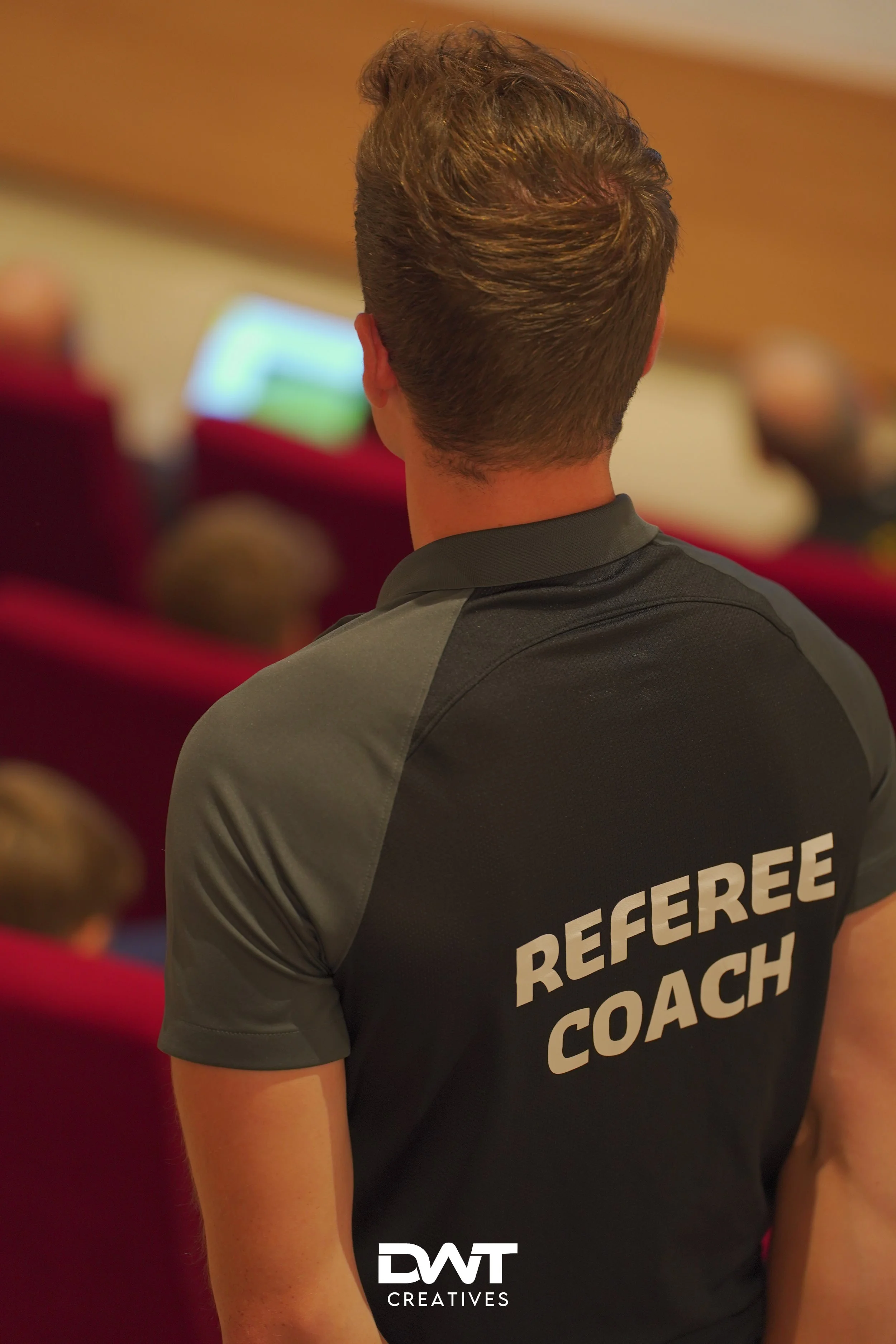 Achteraanzicht van een persoon met een shirt waarop 'Referee Coach' staat geschreven, in een zaal met rode stoelen. De persoon kijkt richting een scherm op de achtergrond.