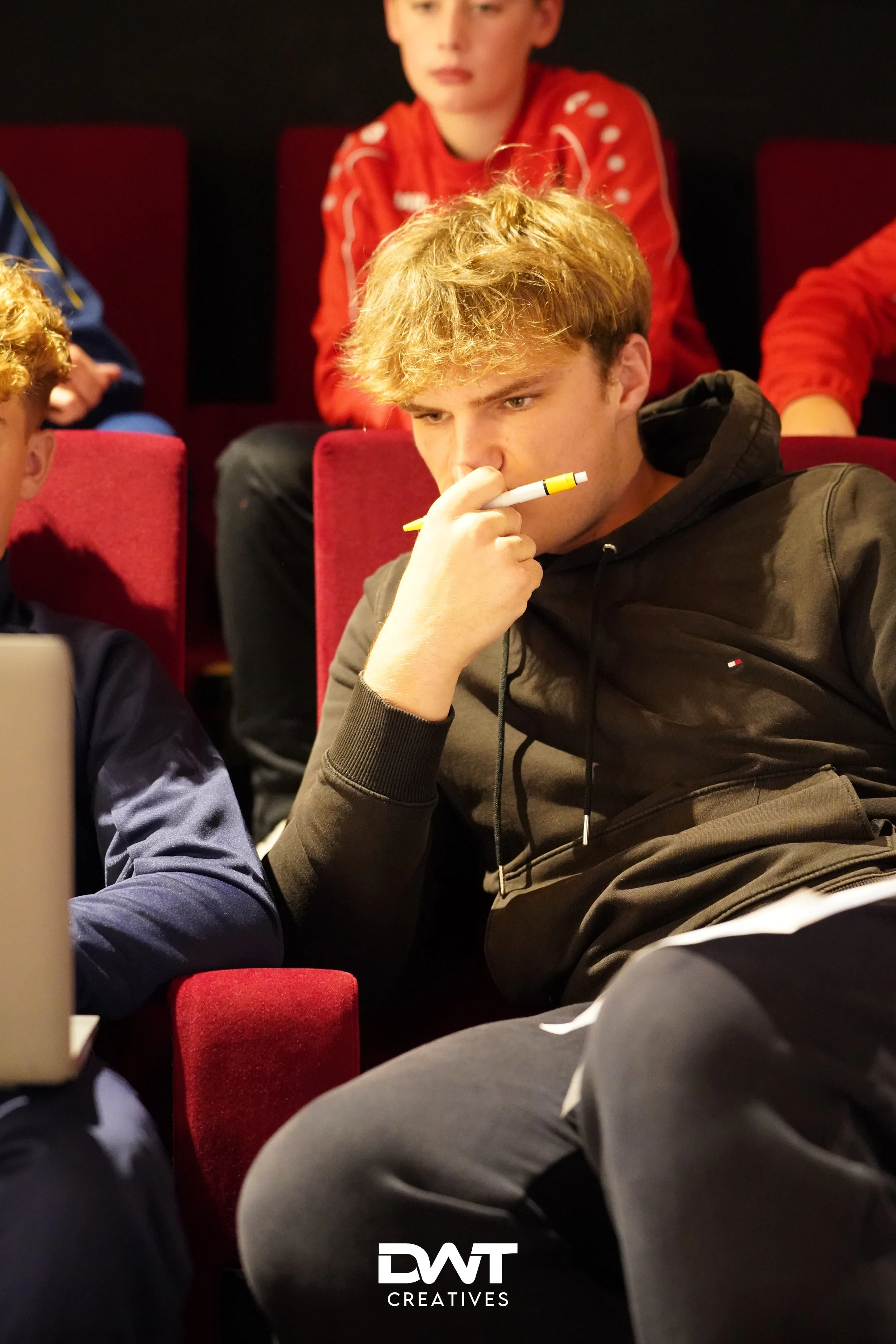 Jonge mannen in een theateropstelling, kijken geconcentreerd naar iets buiten beeld. Een van hen houdt een pen bij zijn gezicht. Het is een sfeervolle setting met gedimde kleuren.