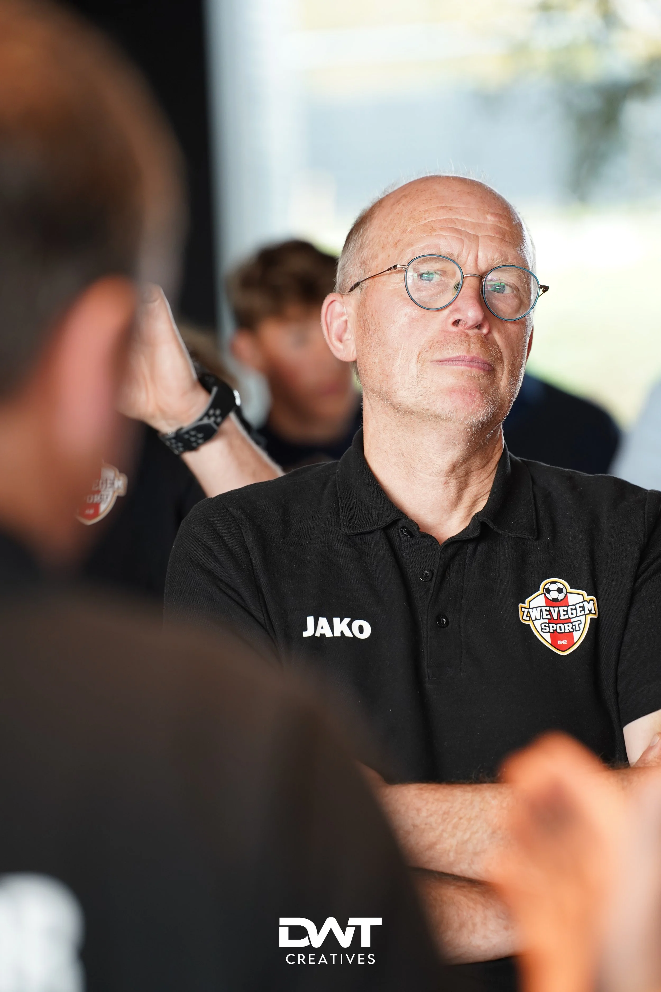 Man met bril en uithangende armen in een zwart T-shirt met "JAKO" en "Zwevegem Sport" embleem. Op de achtergrond zijn andere mensen te zien. Foto door DWT Creatives.