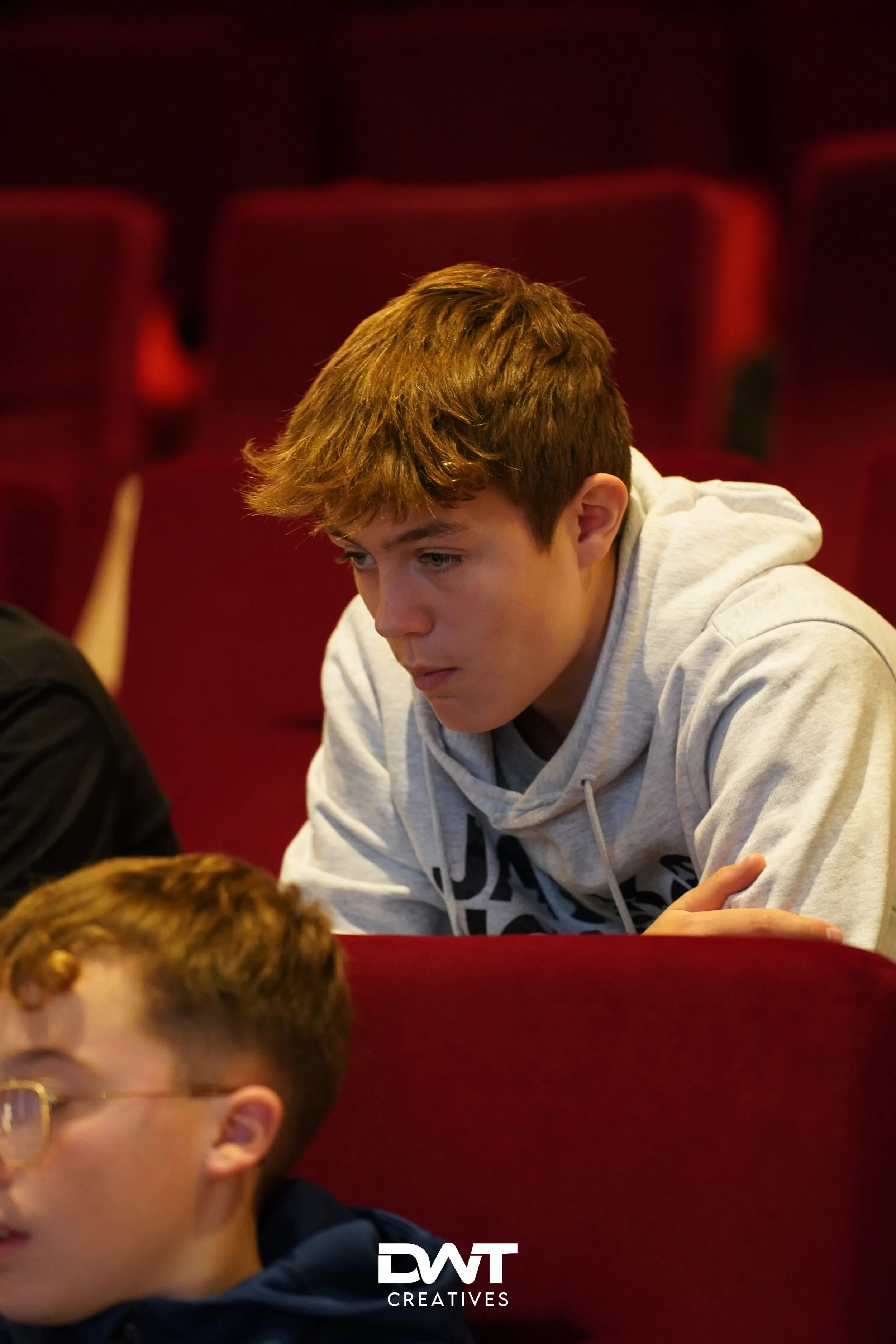 Twee jongens in een theaterzaal, de voorste draagt een bril. Beiden zijn geconcentreerd bezig. Op de achtergrond zijn rode stoelen zichtbaar. Beeldmerk van DWT Creatives.