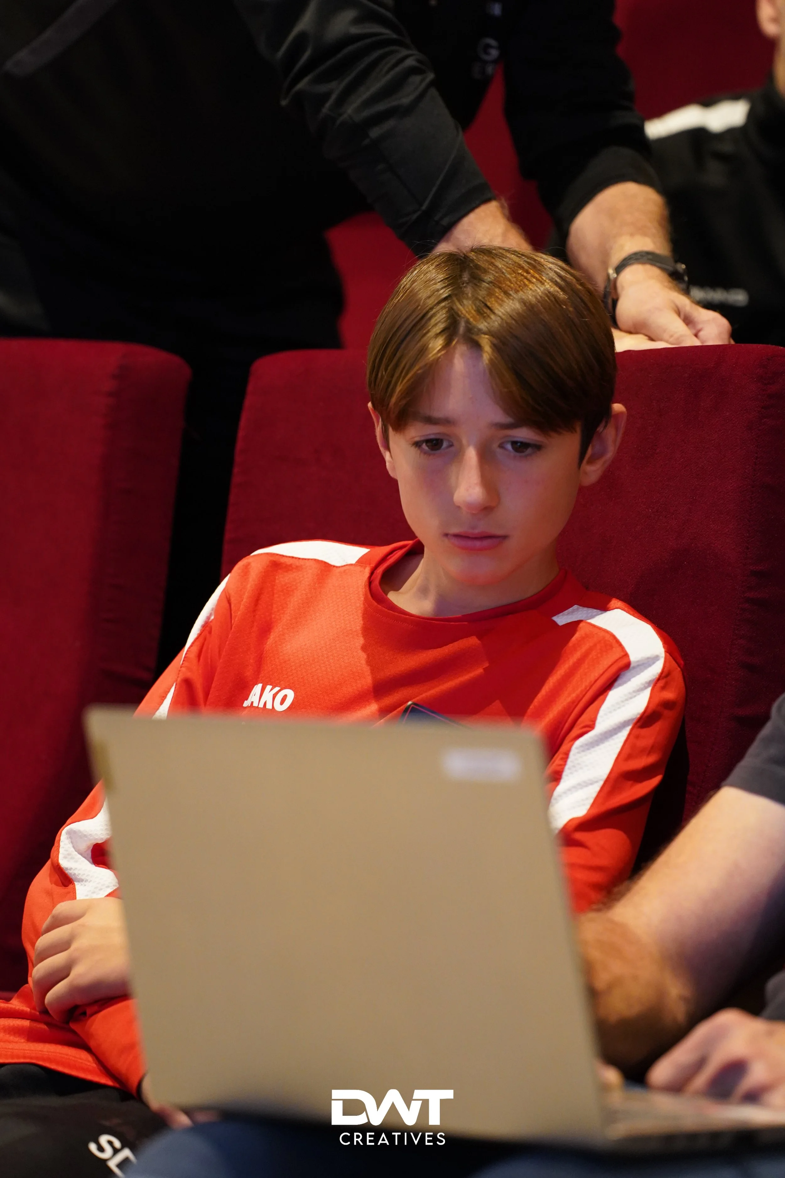 Jongen in rood sportshirt kijkt naar laptop. Hand van een volwassene rust op zijn rug. "DWT Creatives" logo onderaan.