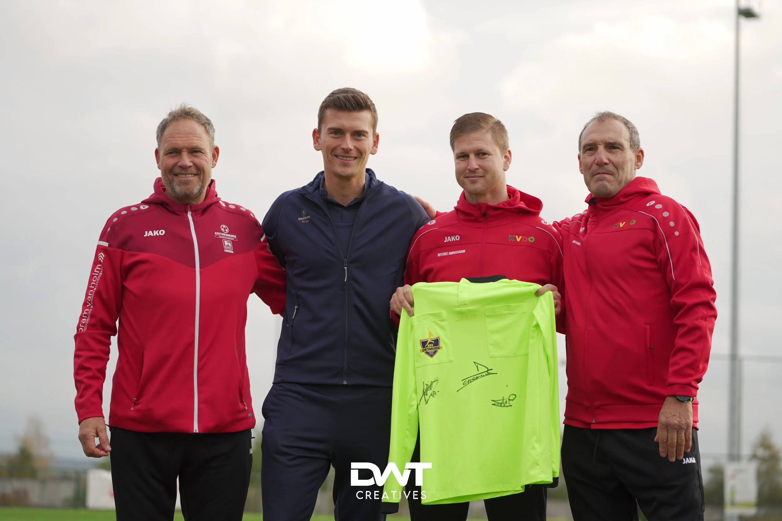Vier mannen poseren voor de camera. Drie dragen rode sportjassen met logo's en merken erop, en de vierde man draagt een donker sportjack. Een van hen houdt een felgeel sportshirt met handtekeningen vast. De mannen staan buiten onder een bewolkte heme