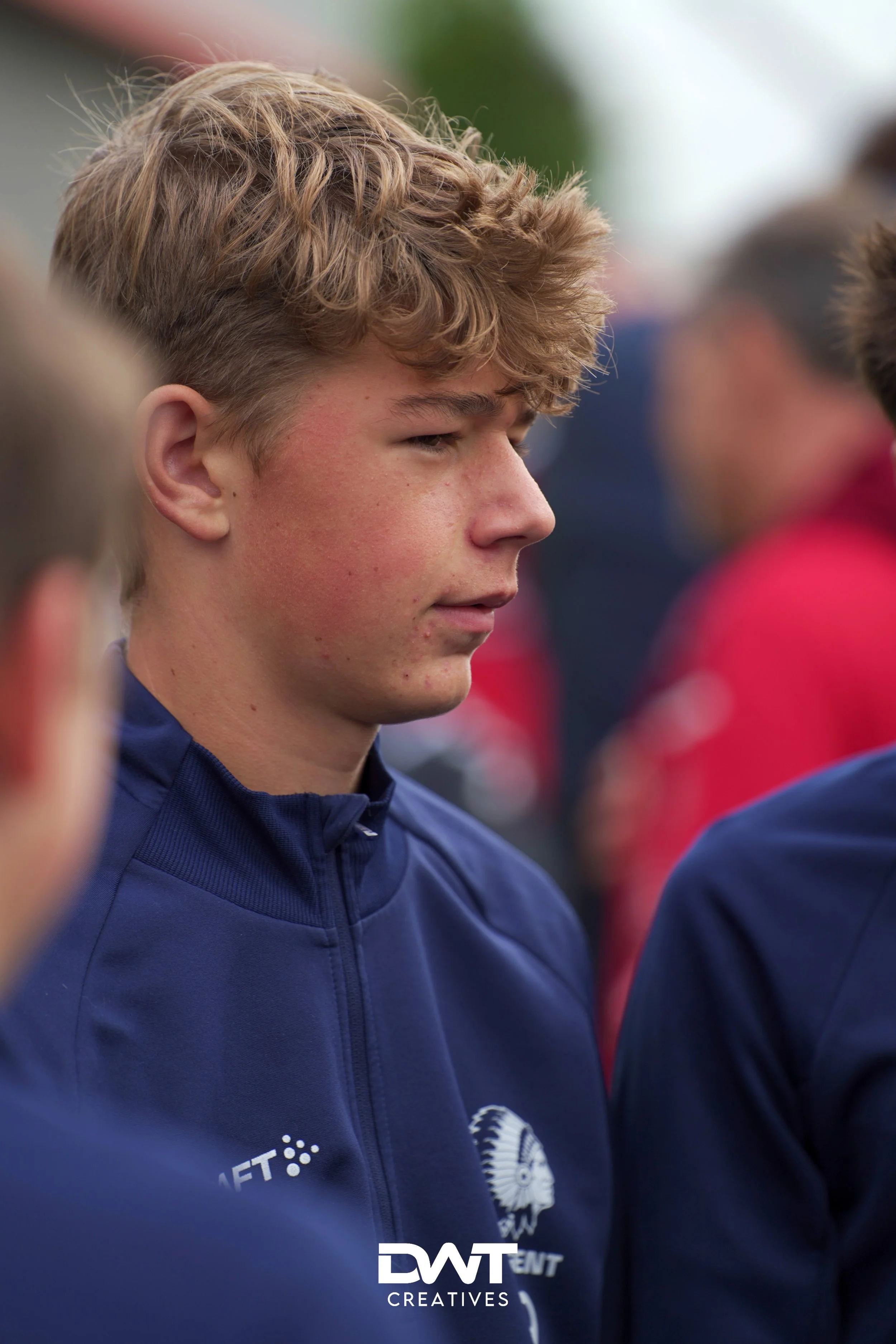 Jonge man in een blauwe jas met een sportlogo, buiten in een groepsomgeving.