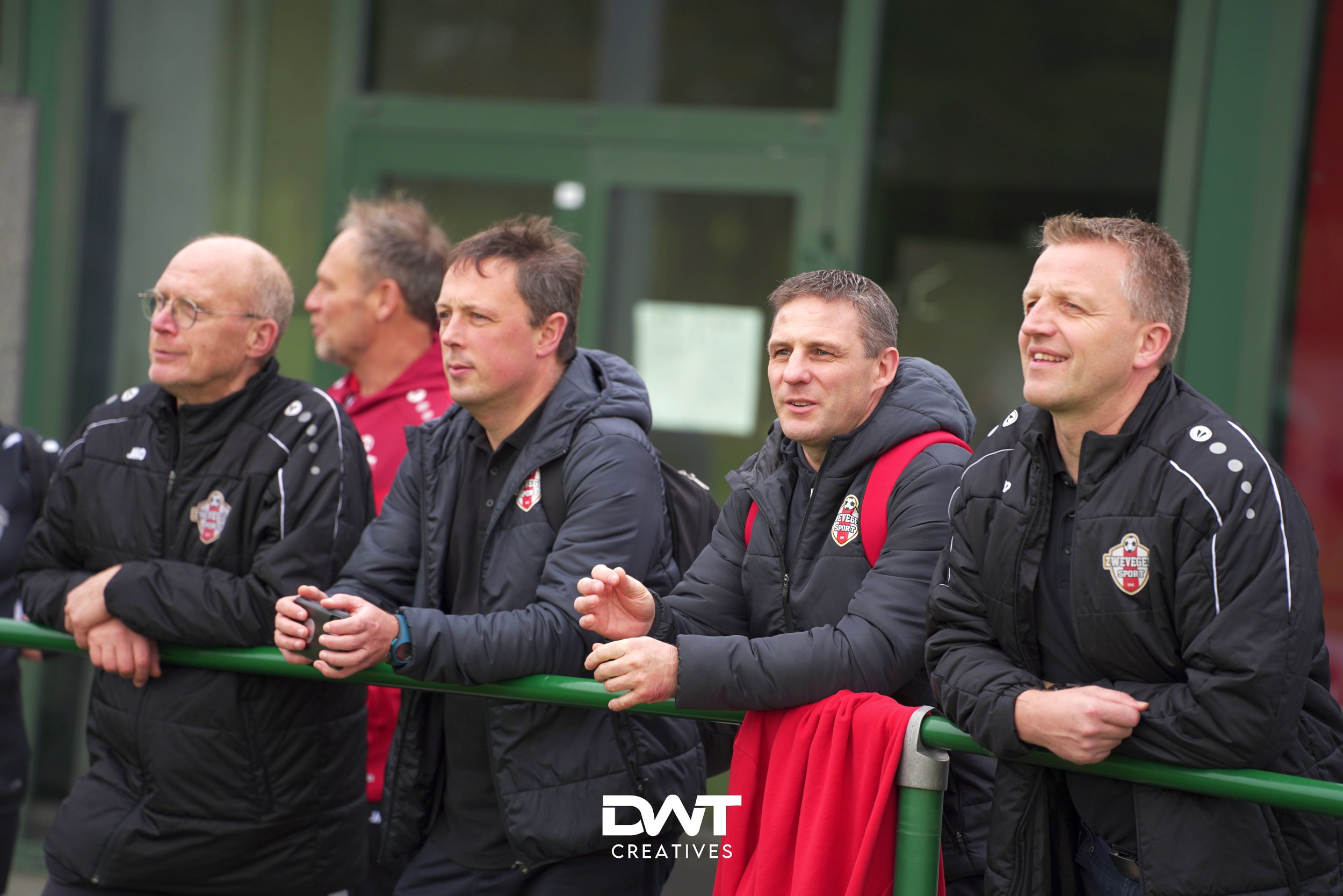 Groep mannen in sportjassen kijkt aandachtig naar een sportevenement op een voetbalveld. Ze dragen jassen met een clublogo.