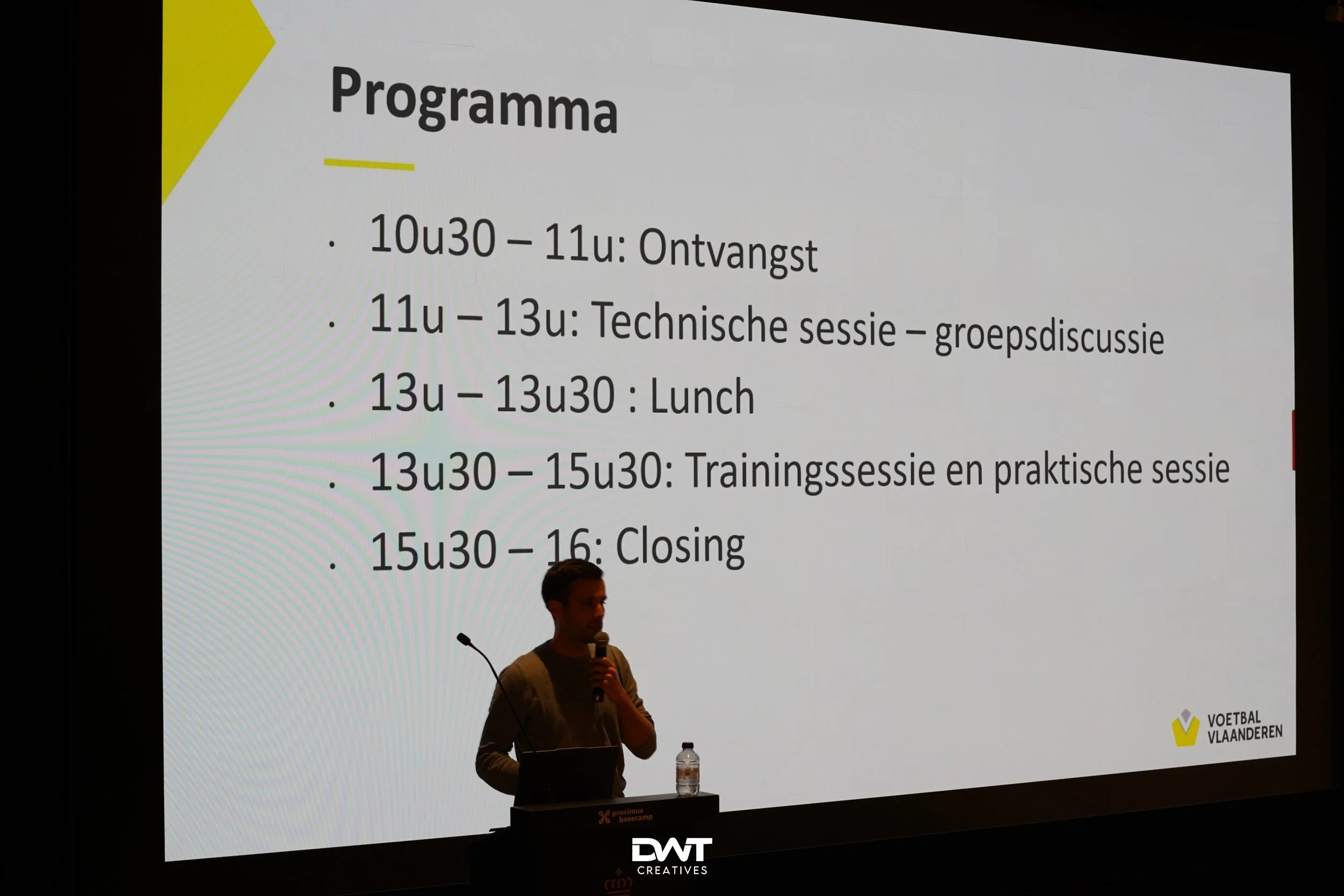 Een man met een microfoon staat voor een scherm met een programma. Het programma omvat verschillende sessies: ontvangst, technische sessie, lunch, trainingssessie en afsluiting, georganiseerd door Voetbal Vlaanderen.
