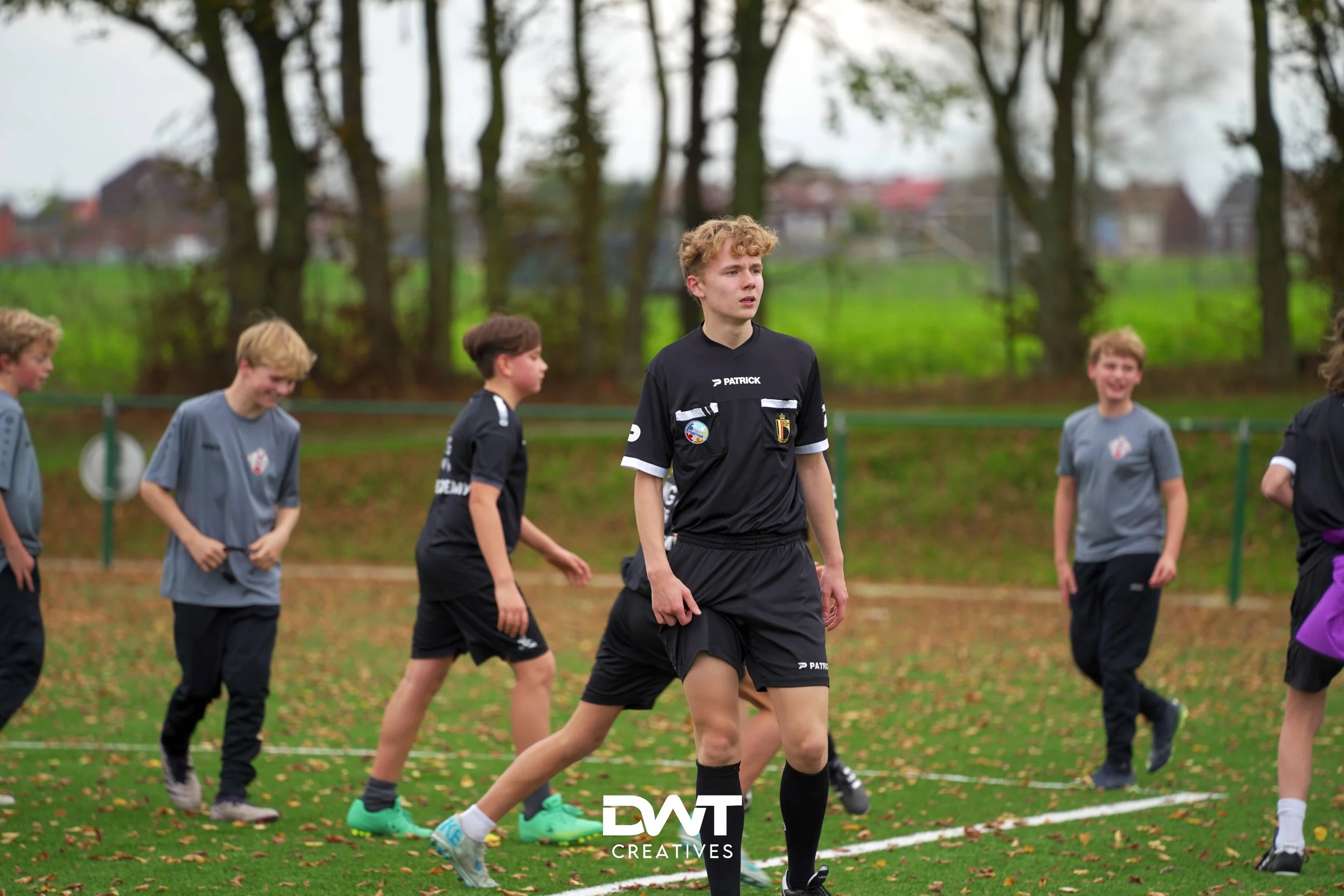 Jongens spelen voetbal op een grasveld in een park, een draagt een zwart tenue met gele accenten, anderen dragen grijze shirts. Er zijn bomen op de achtergrond.