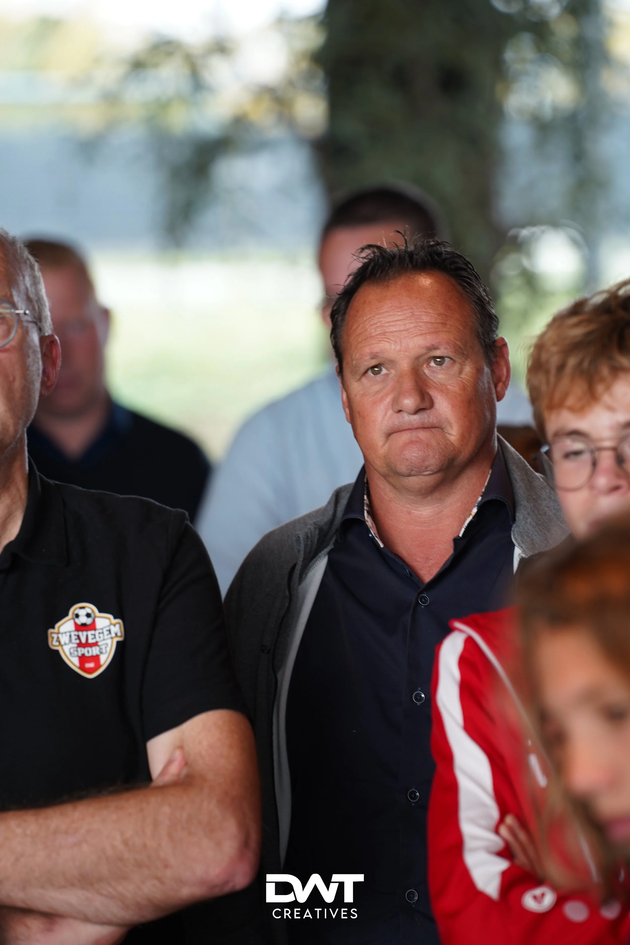 Groep mensen kijkt aandachtig bij een sportevenement, man links draagt T-shirt met "Zwevegem Sport" logo. Achtergrond is onscherp.