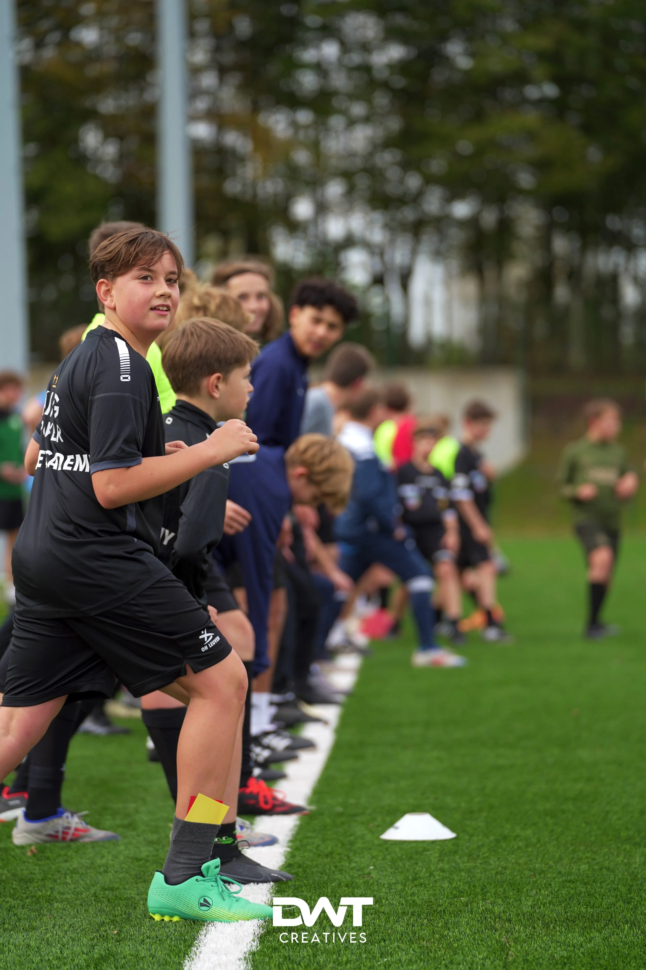 Kinderen staan in een rij op een grasveld klaar voor een activiteit of oefening, mogelijk op een sportveld. Ze dragen sportkleding en kijken geconcentreerd naar voren. "DWT CREATIVES" logo onderaan de foto.