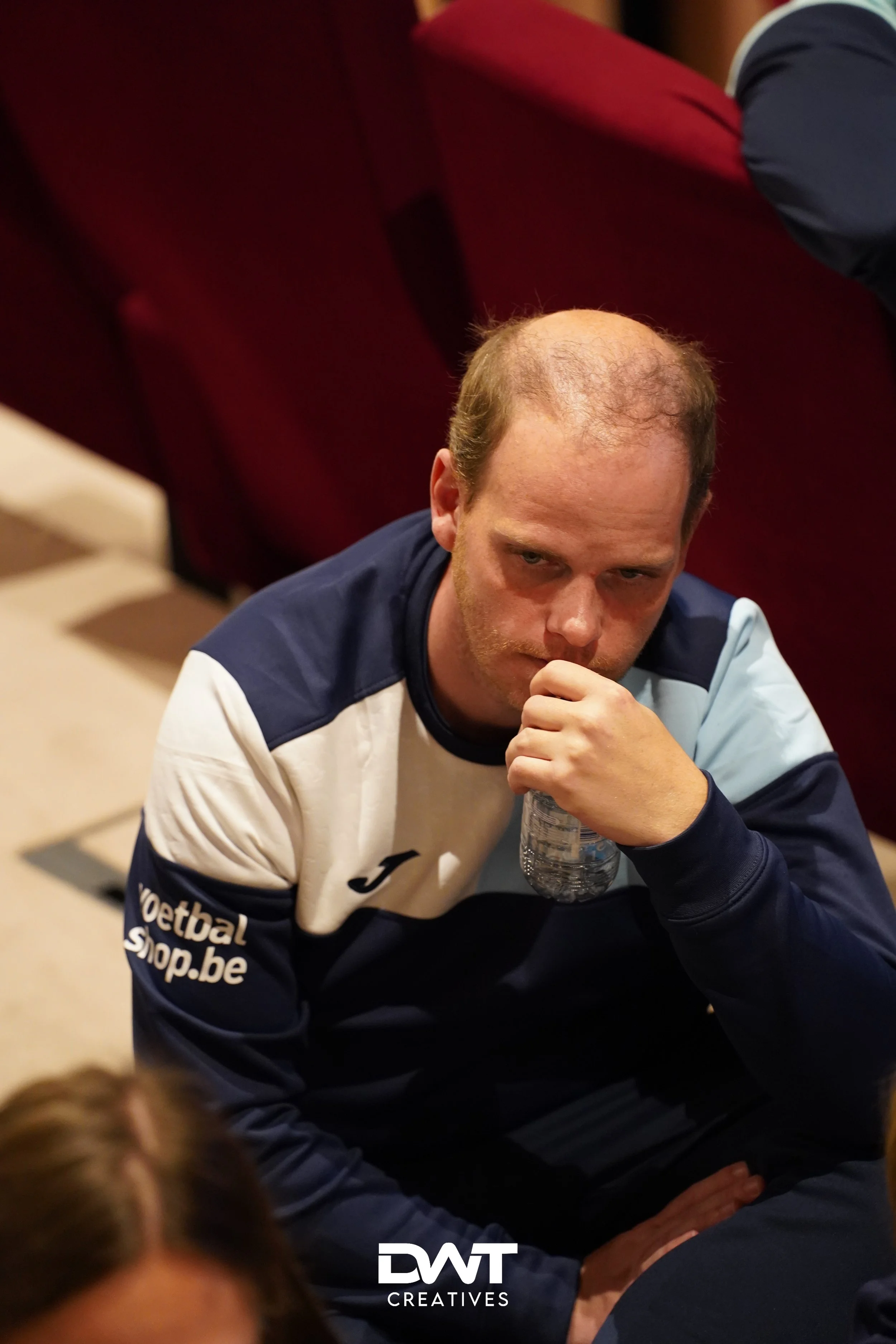 Man zittend met hand aan mond en waterfles, gekleed in sportkleding, in een ruimte met rode stoelen.