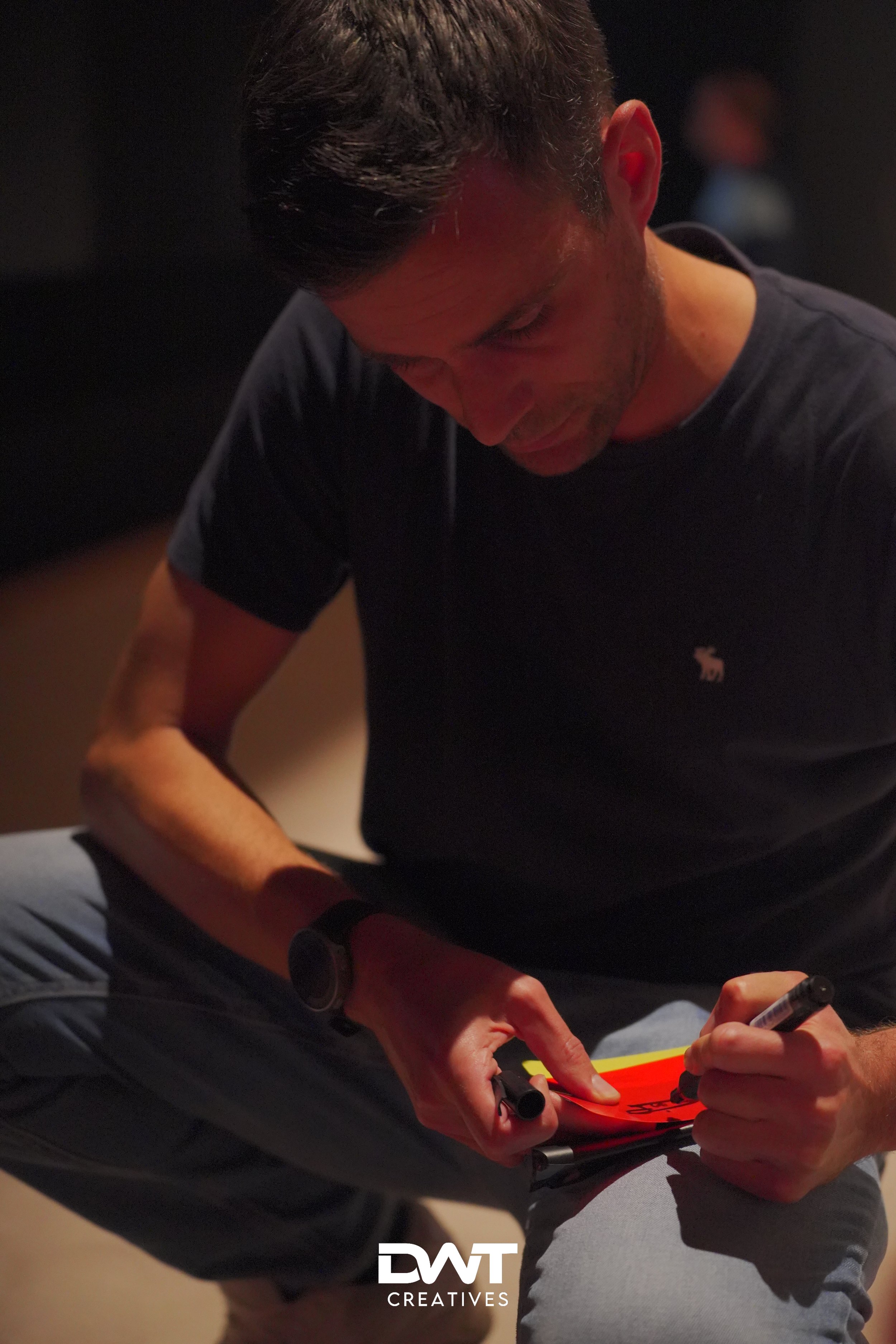 Een man in een donker shirt schrijft op een oranje notitieblok met een stift, terwijl hij gehurkt zit. De scène is in een donkere omgeving.