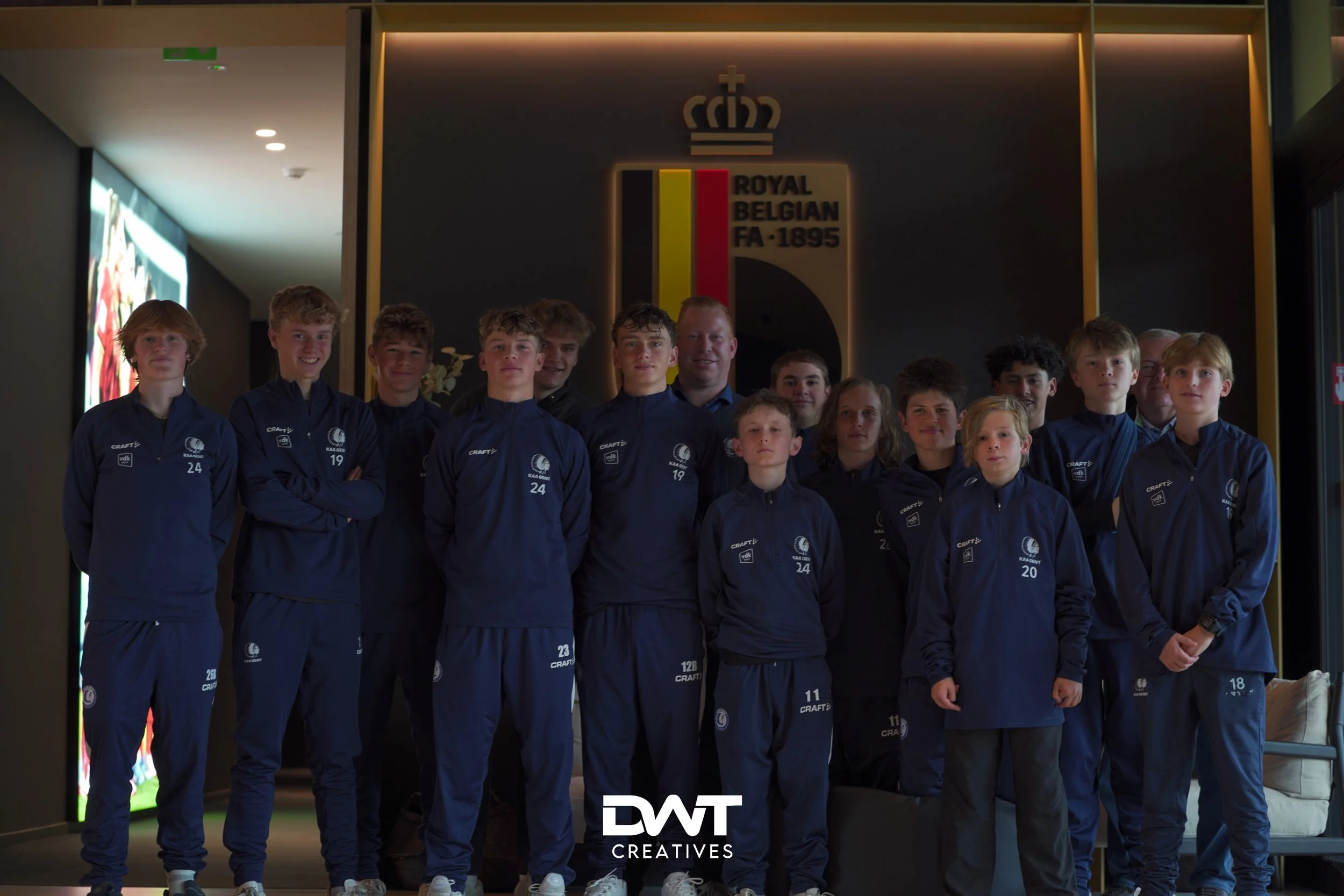 Groep jonge voetballers in trainingspakken poseren voor een bord met 'Royal Belgian FA - 1895' logo.